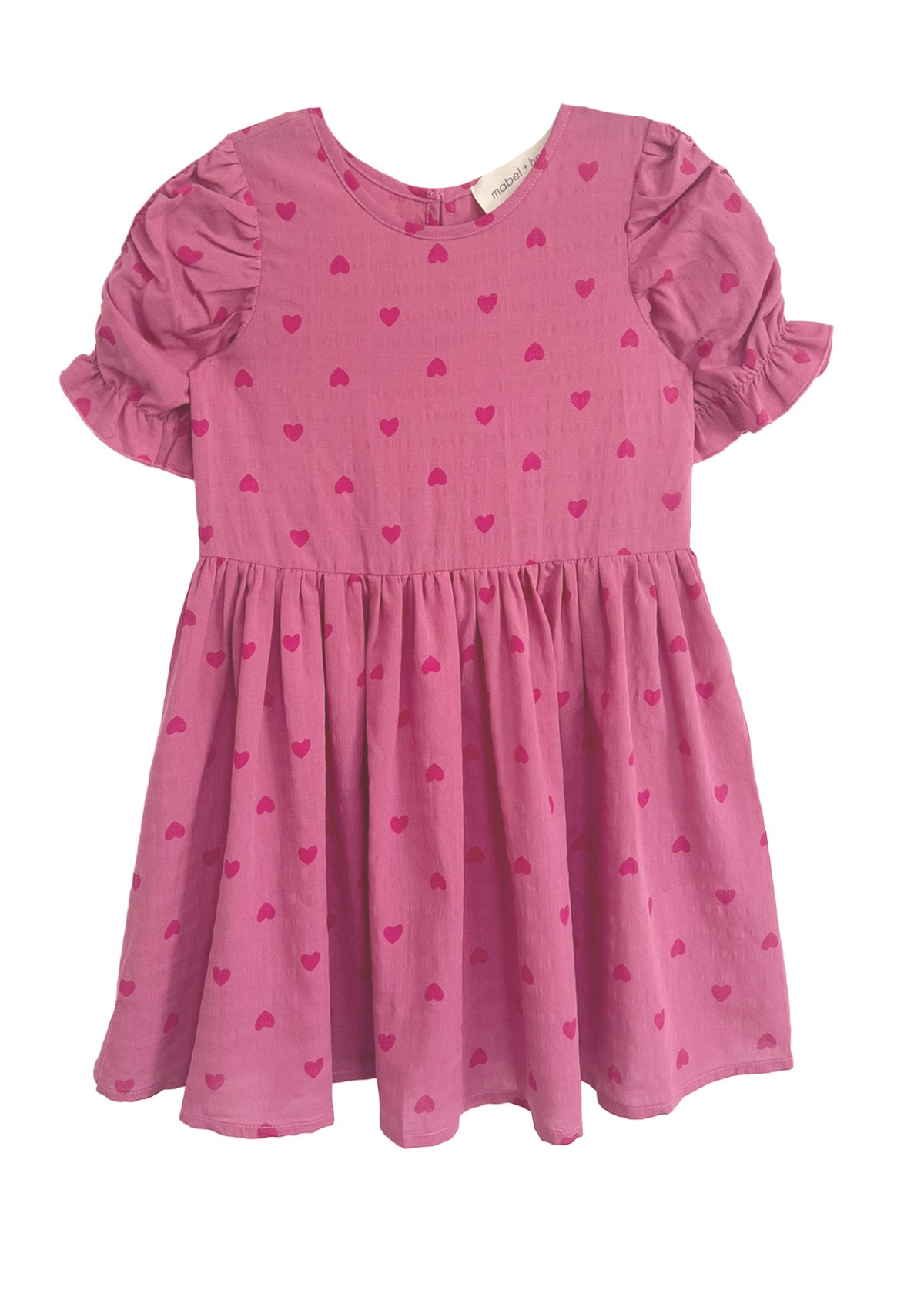The Mabel and Honey Perfect in Pink Hearts Short Sleeve Dress is a girls’ pink dress with puffed ruffled sleeves, a flared skirt, and a sweet pattern of small, darker pink hearts.