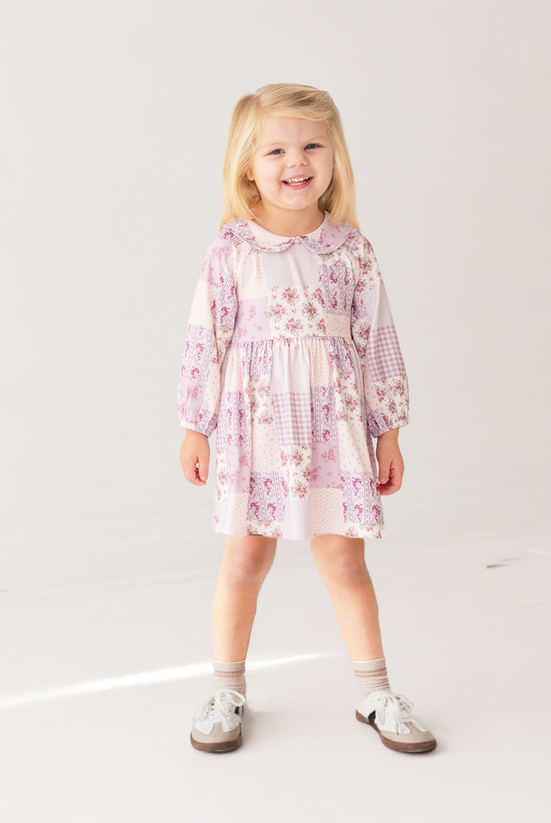 A young girl with blonde hair smiles while wearing the Liliana Floral Dress by Mabel and Honey—featuring long sleeves, a pink and white pattern, and a Peter Pan collar—paired with white socks and brown shoes against a white background.