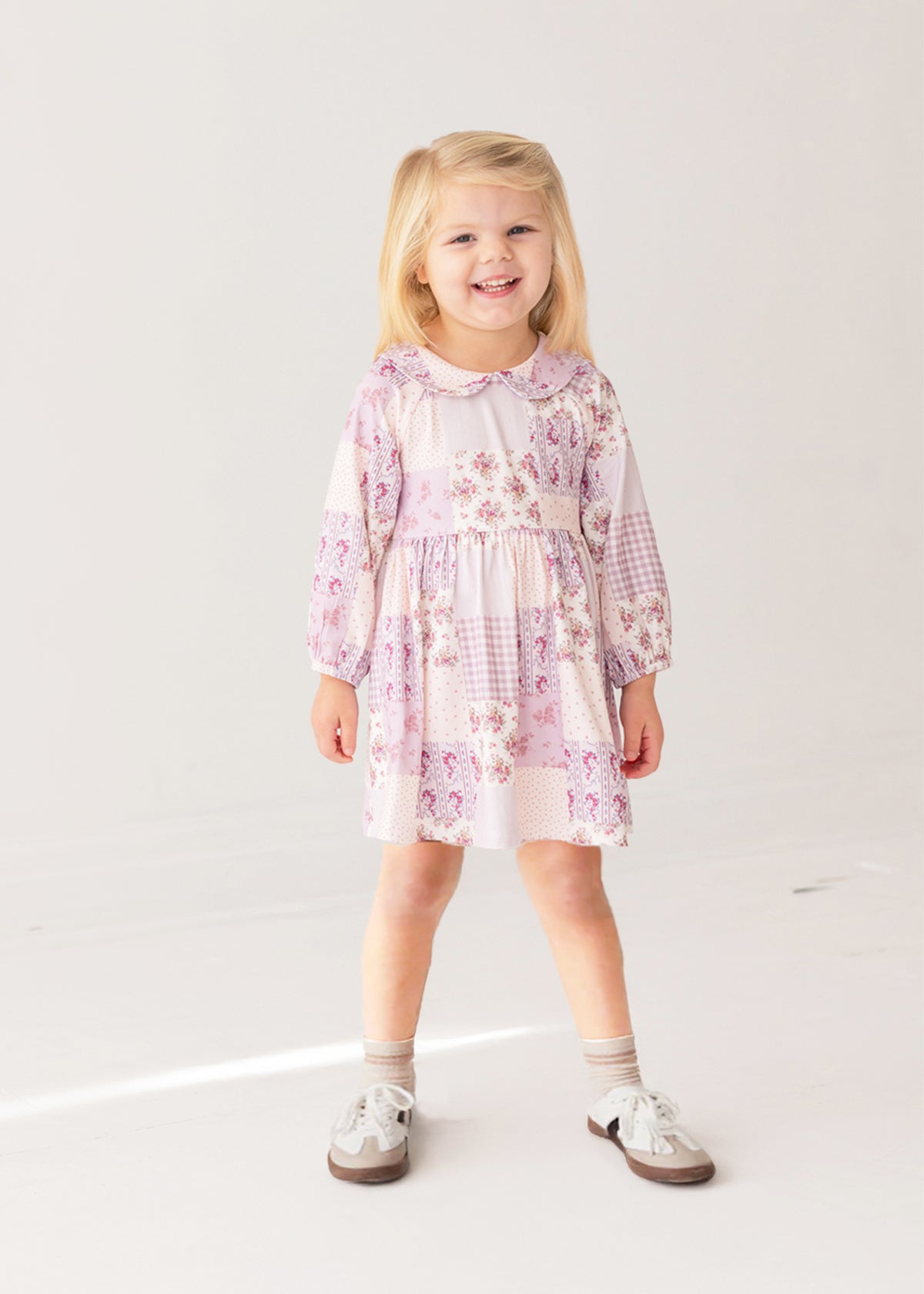 A young girl with blonde hair smiles while wearing the Liliana Floral Dress by Mabel and Honey—featuring long sleeves, a pink and white pattern, and a Peter Pan collar—paired with white socks and brown shoes against a white background.