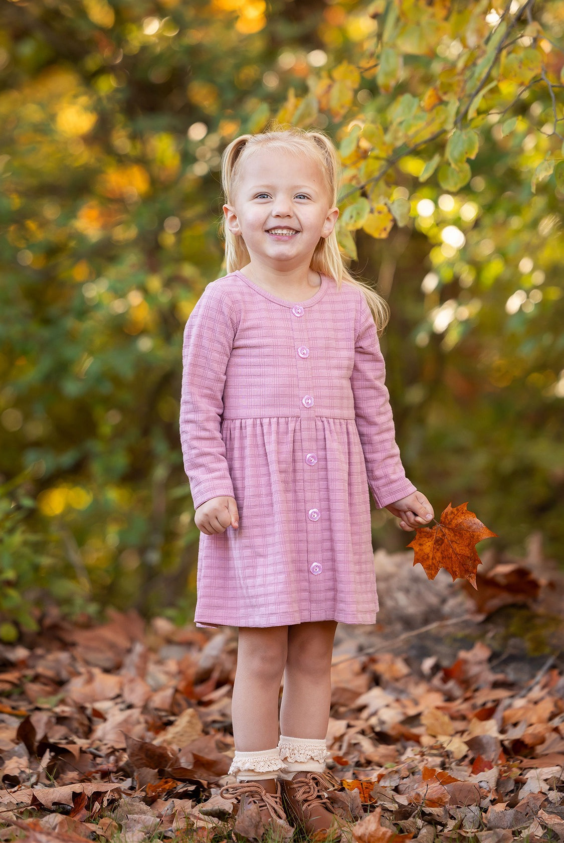 A young girl with blonde pigtails smiles among fallen autumn leaves, holding an orange leaf. She wears a Mabel and Honey Liliana Plaid Dress with green and yellow foliage in the background.