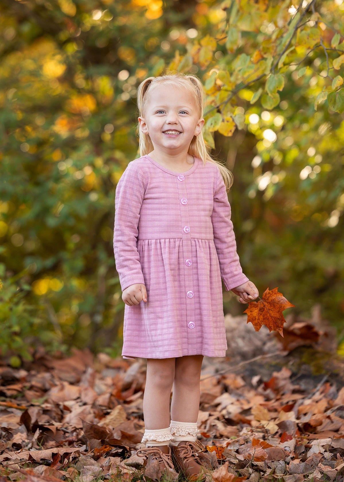 A young girl with blonde pigtails smiles among fallen autumn leaves, holding an orange leaf. She wears a Mabel and Honey Liliana Plaid Dress with green and yellow foliage in the background.