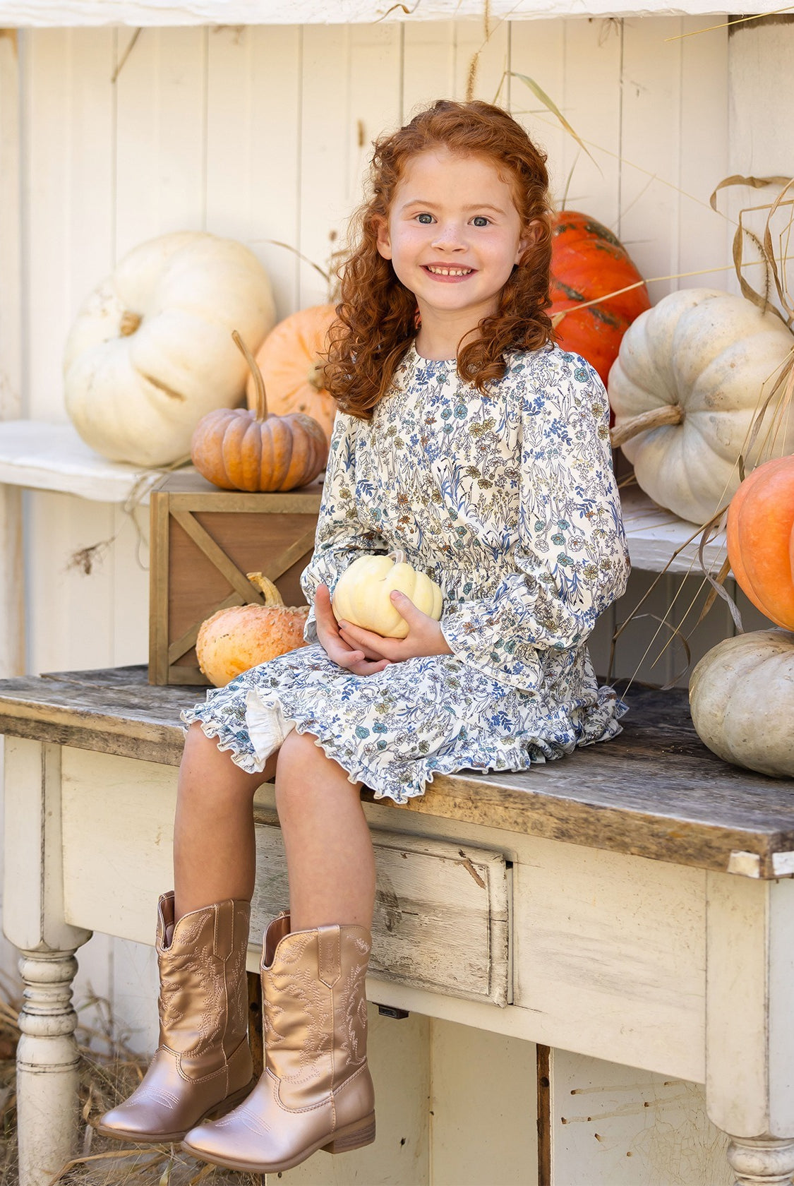 A young girl with curly red hair, wearing the Mabel and Honey Brooklyn Floral Dress and brown boots, sits on a rustic bench holding a small white pumpkin. She smiles, surrounded by white and orange pumpkins against a white wooden backdrop.