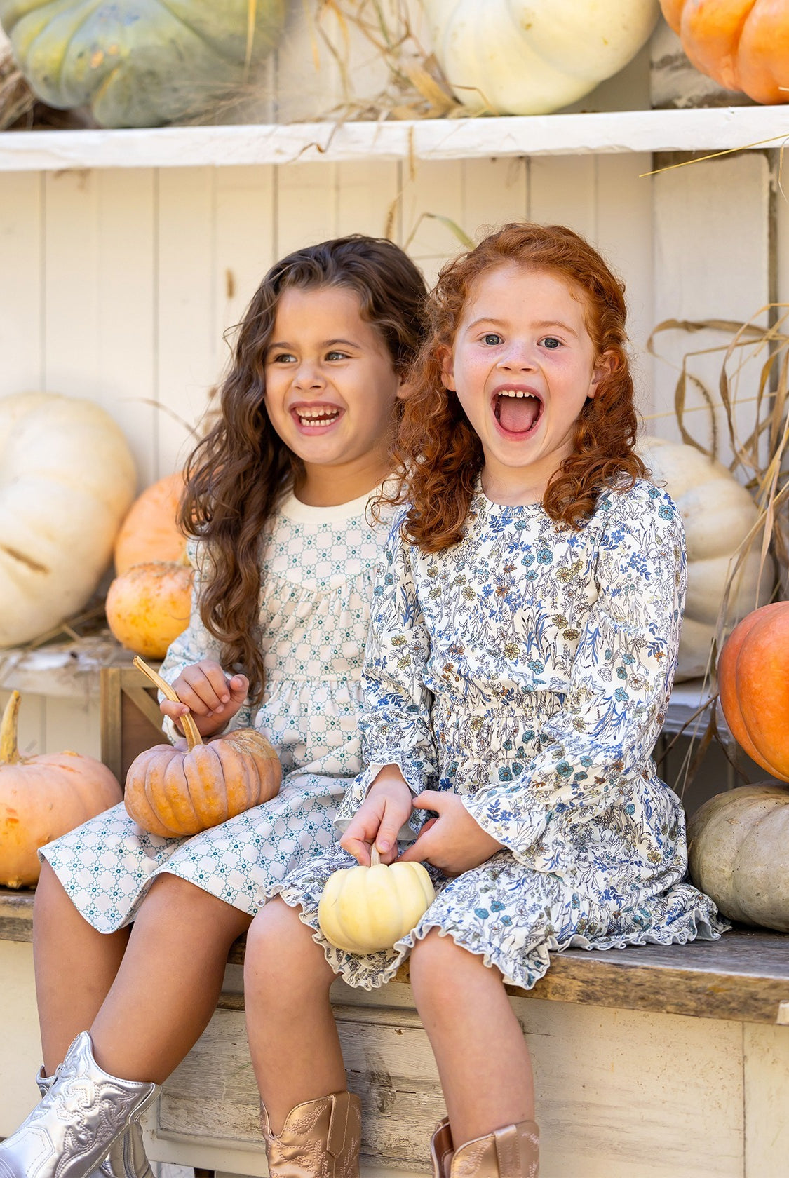 Two young girls in Mabel and Honey's Brooklyn Floral Dress sit on a bench, smiling with small pumpkins. Metallic boots and shelves of white and orange pumpkins behind them create a festive autumn scene.