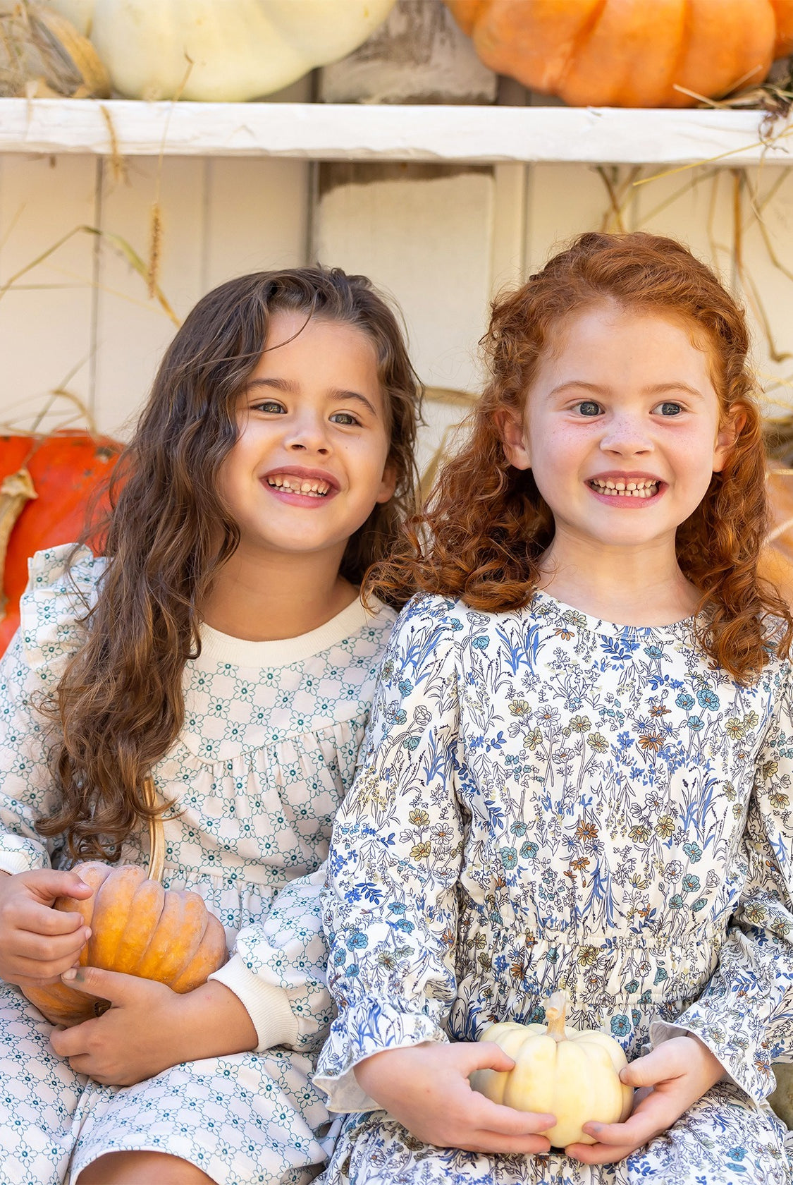 Two young girls smile brightly in their Brooklyn Floral Dresses by Mabel and Honey, each holding a small pumpkin. Orange and white pumpkins in the background add a warm autumn touch to the scene.
