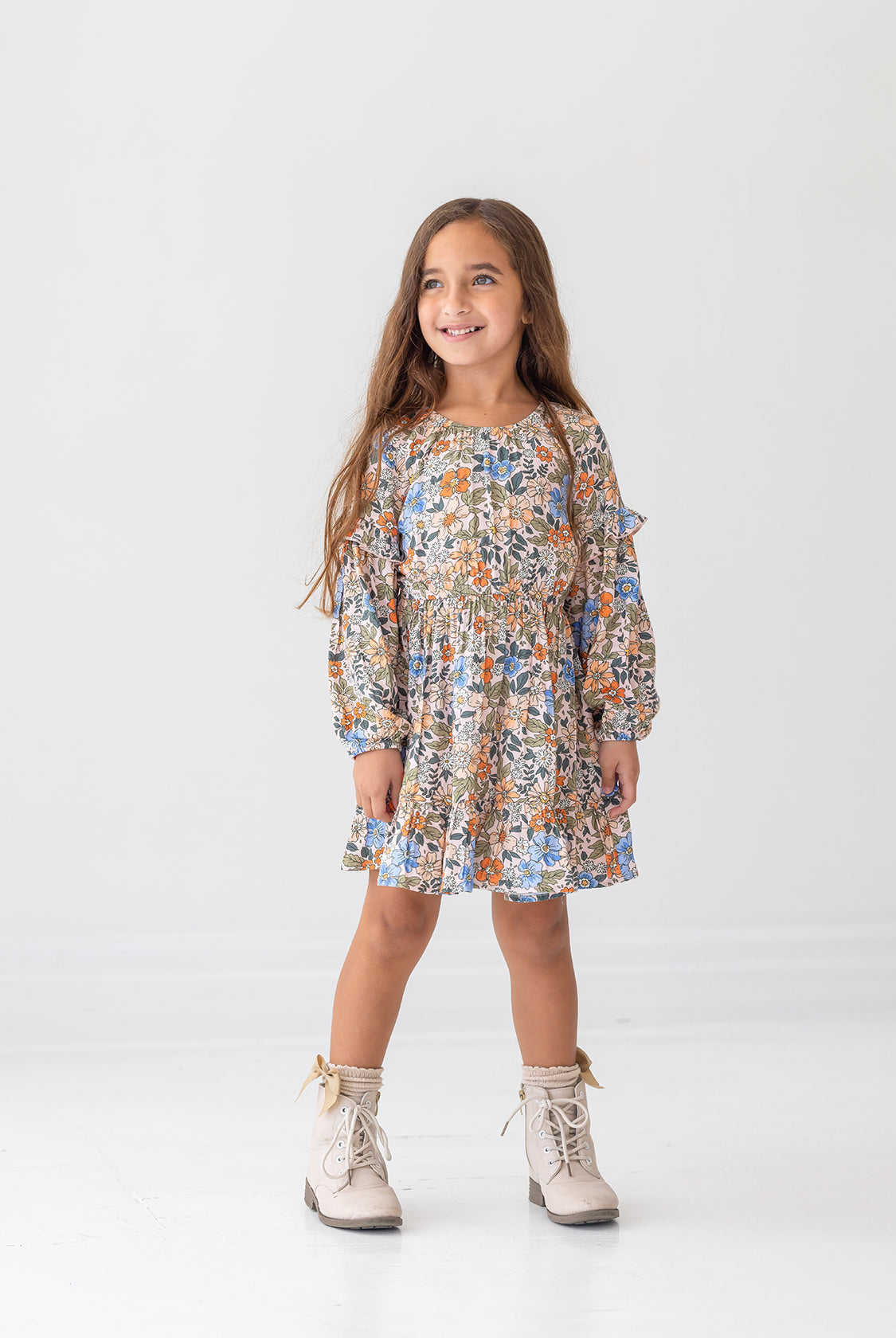 A young girl with long brown hair smiles against a white background, wearing the Mabel and Honey Riley Long Sleeve Dress with a floral print and beige lace-up boots.