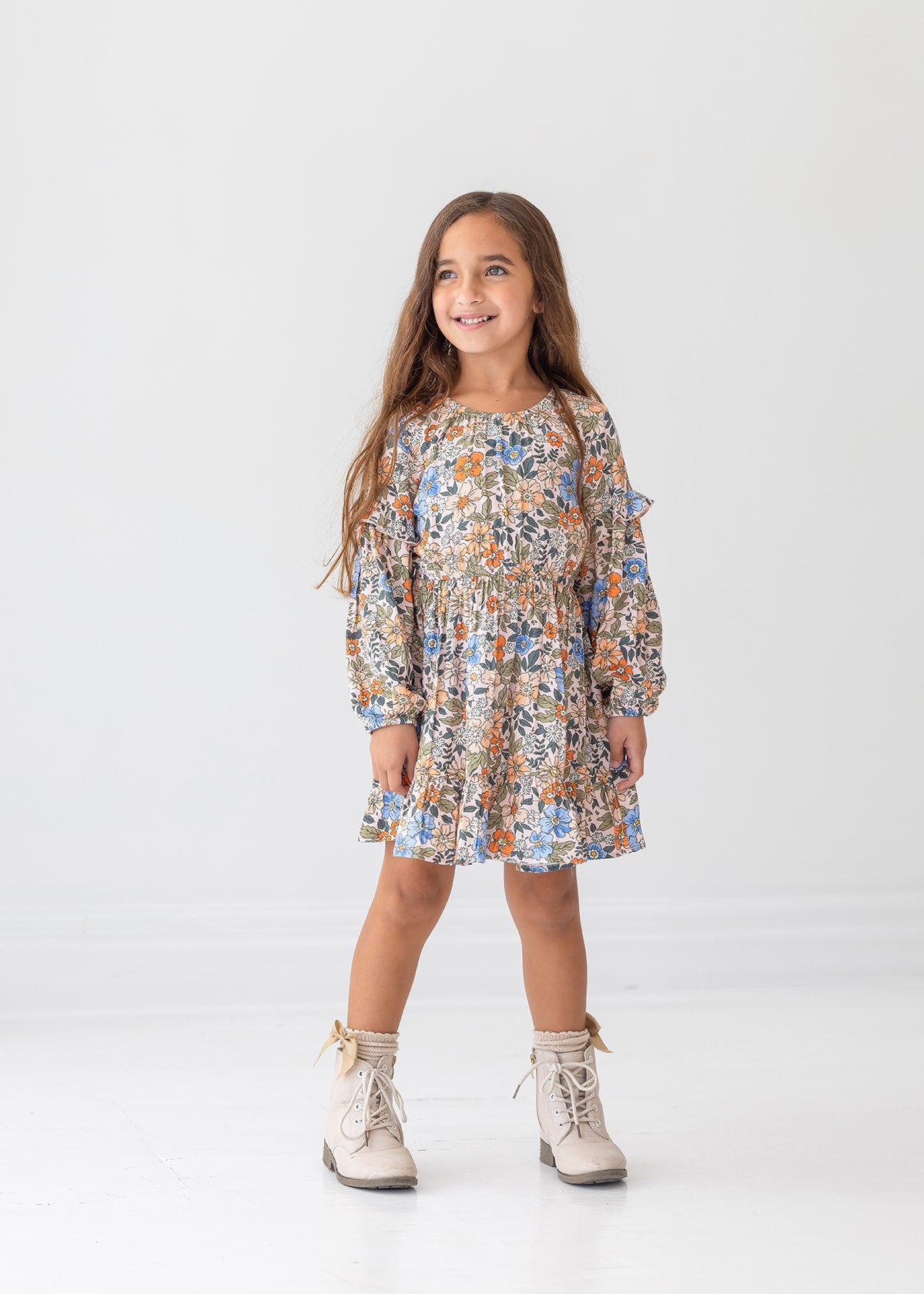 A young girl with long brown hair smiles against a white background, wearing the Mabel and Honey Riley Long Sleeve Dress with a floral print and beige lace-up boots.