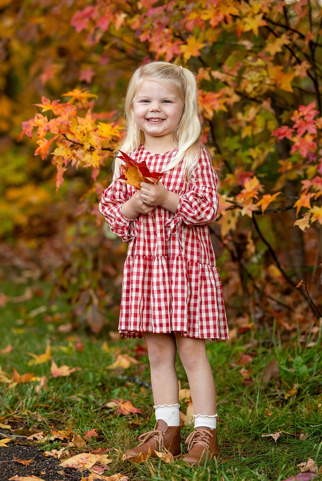 A young girl smiles outdoors, holding orange autumn leaves and wearing the Mabel and Honey Tis' the Season Dress, surrounded by colorful fall foliage.