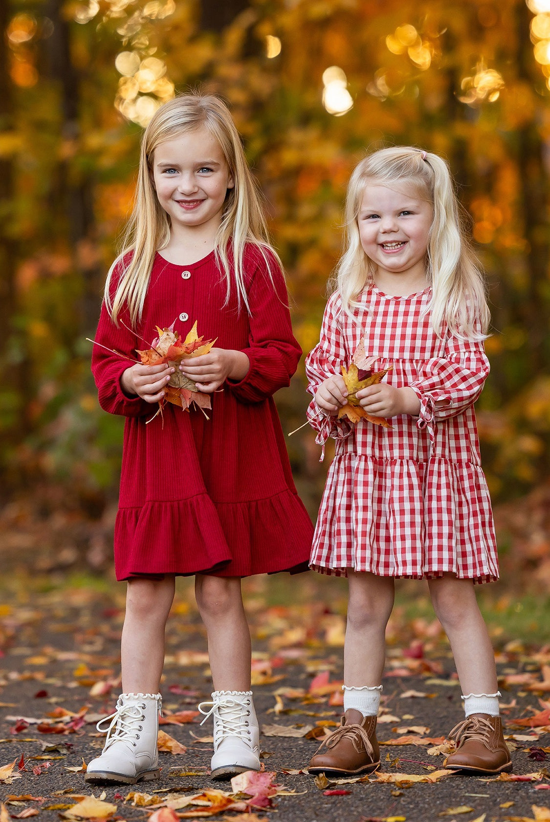 Two girls smile outdoors among fall foliage. One wears the Mabel and Honey Tis' the Season Dress with white boots, while the other sports a red gingham dress and brown shoes. Both hold autumn leaves in their hands.
