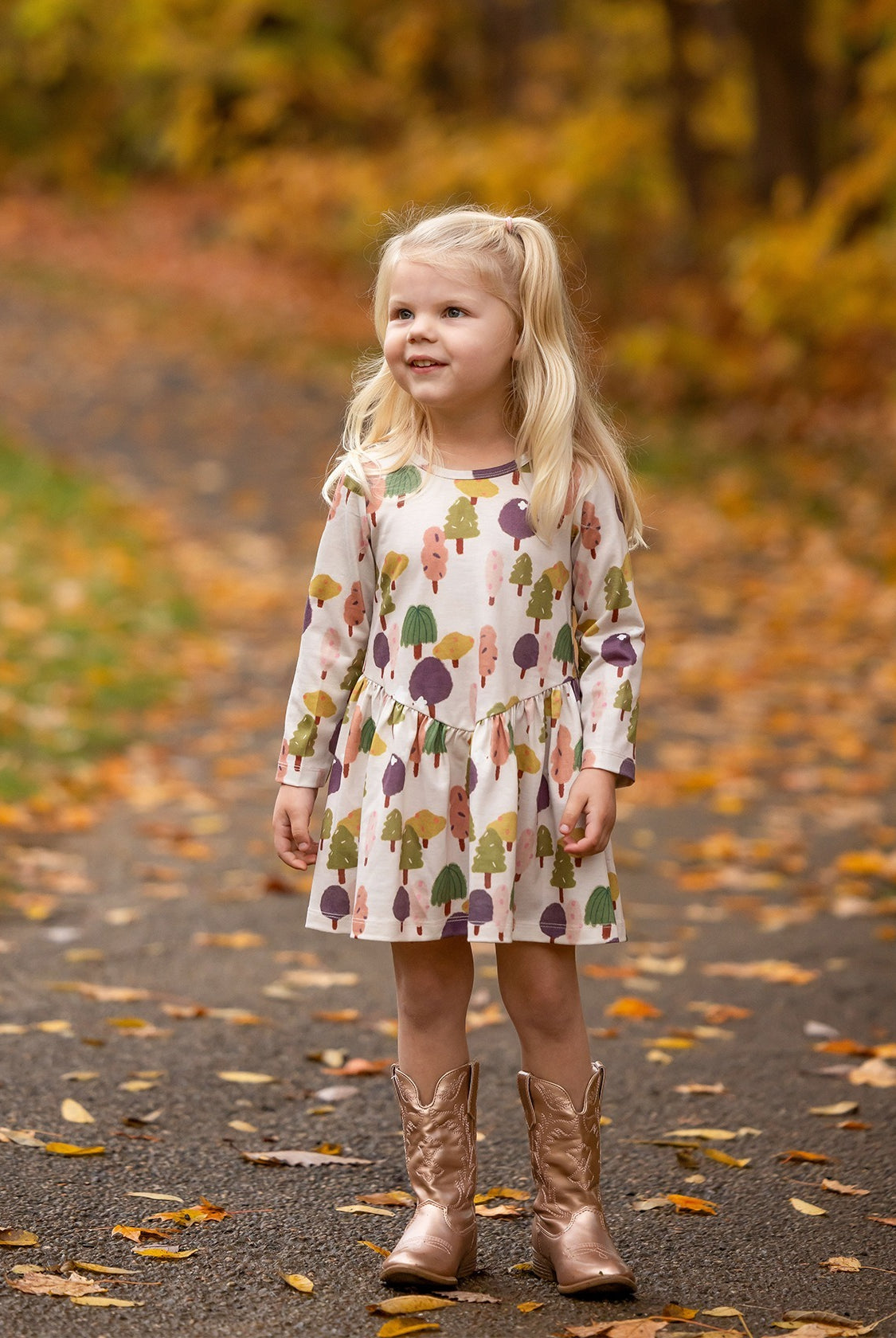 A young girl with long blonde hair smiles on a leafy autumn path, wearing the Treehouse Dress by Mabel and Honey. The nature-inspired style adds playful elegance as she blends into the golden fall scenery.