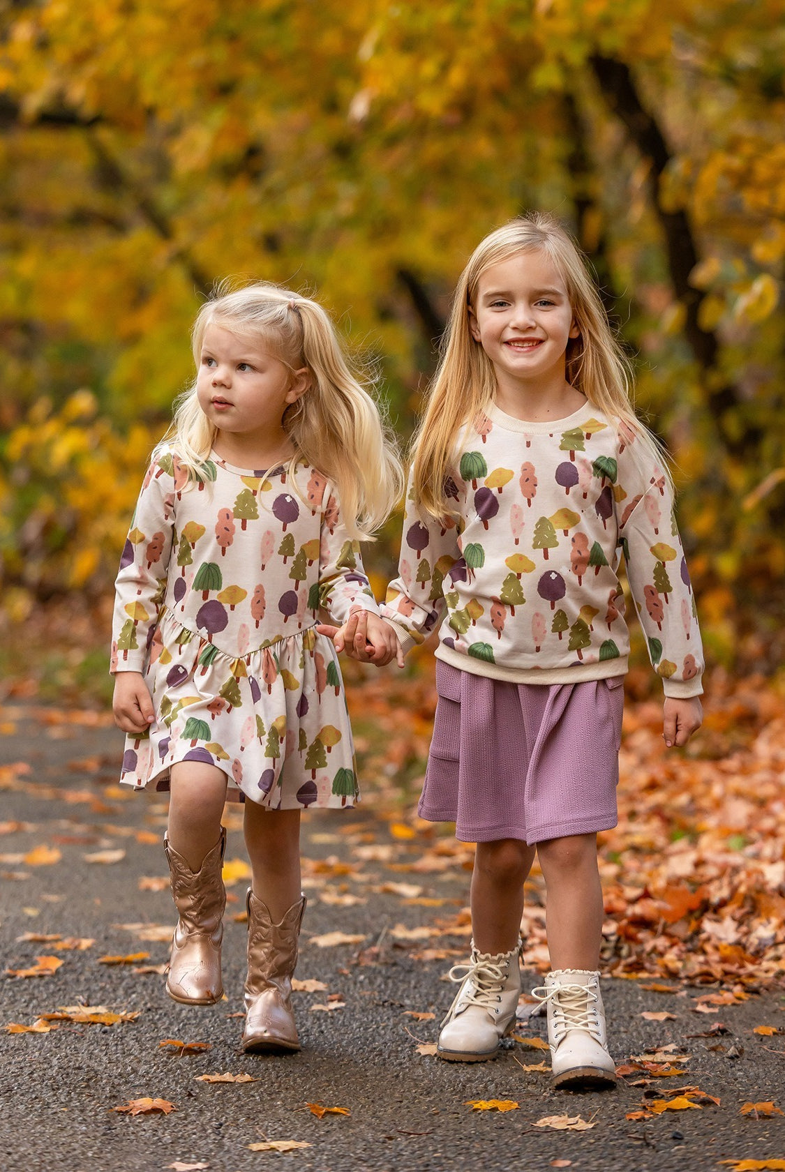 Two young girls stroll hand in hand on a leafy autumn path, dressed in matching Mabel and Honey Treehouse Dresses. One smiles at the camera as they’re surrounded by golden fall foliage and nature-inspired style.