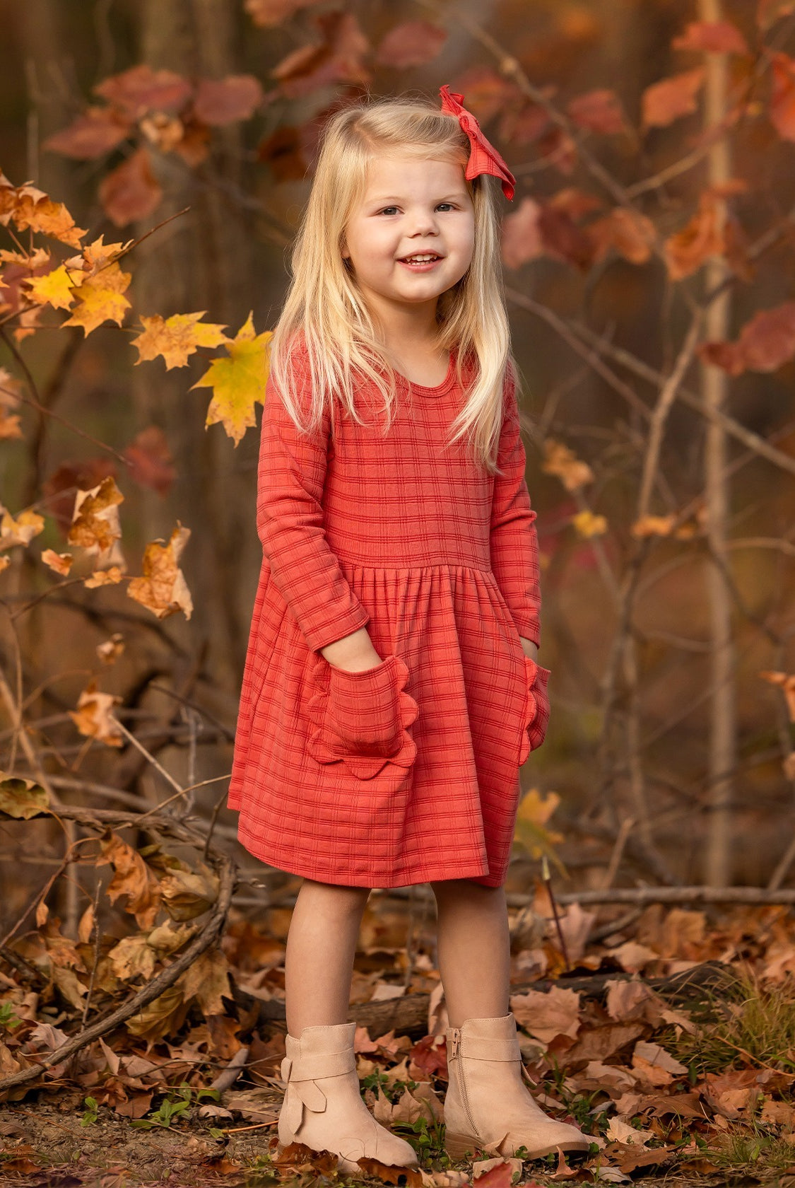 A young girl with long blonde hair, wearing the Mabel and Honey Tea Party Red Gingham Dress and tan boots, stands smiling outdoors among autumn leaves with her hands in her pockets—capturing everyday elegance.