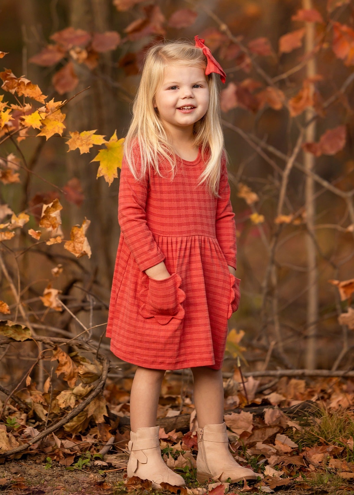 A young girl with long blonde hair, wearing the Mabel and Honey Tea Party Red Gingham Dress and tan boots, stands smiling outdoors among autumn leaves with her hands in her pockets—capturing everyday elegance.