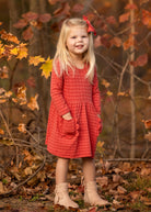 A young girl with long blonde hair, wearing the Mabel and Honey Tea Party Red Gingham Dress and tan boots, stands smiling outdoors among autumn leaves with her hands in her pockets—capturing everyday elegance.