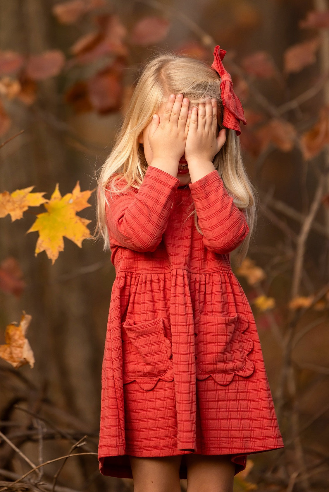 A young girl with long blonde hair, wearing the Mabel and Honey Tea Party Red Gingham Dress and a matching bow, stands outdoors by autumn leaves, shyly covering her face—a sweet display of everyday elegance.