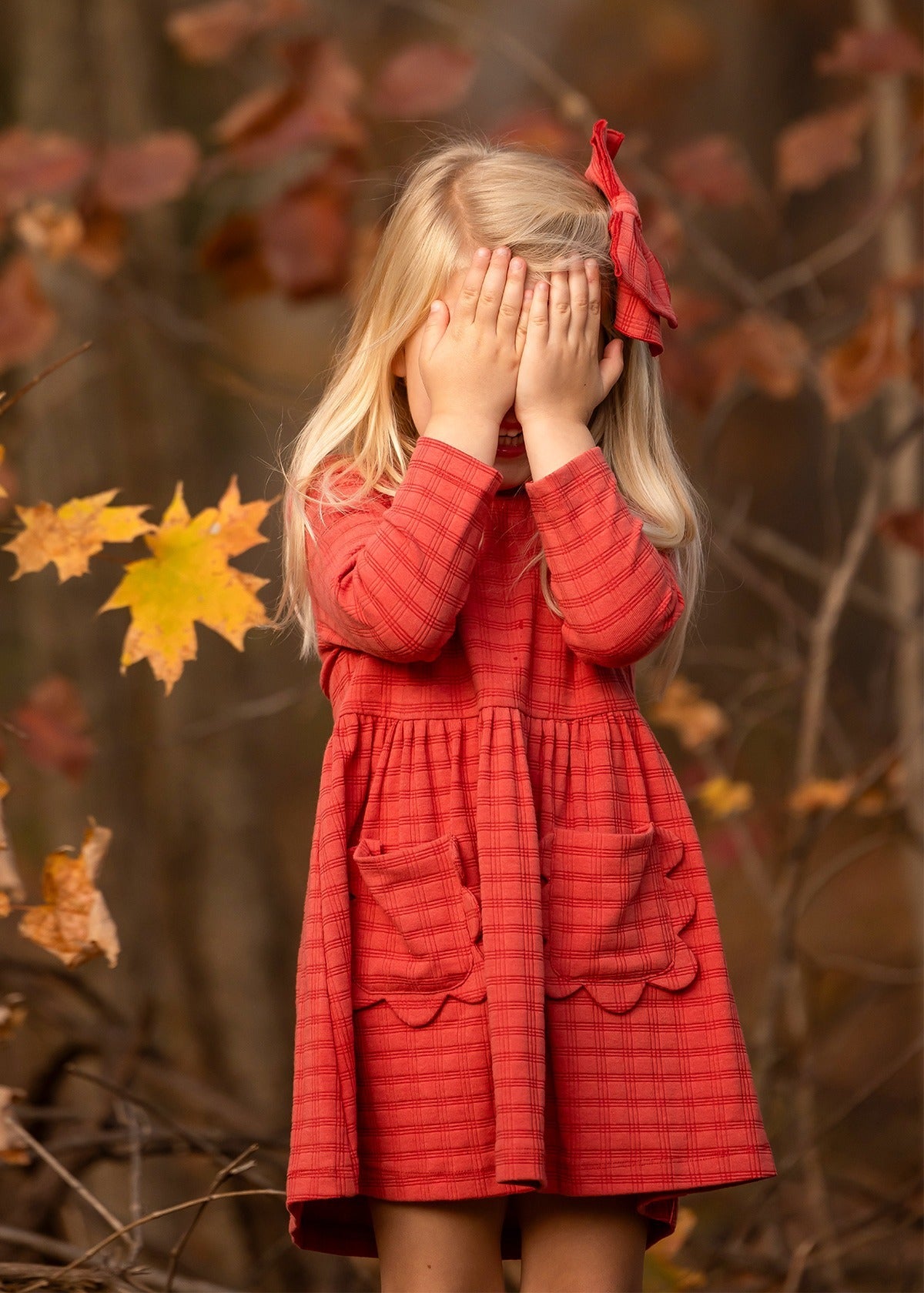 A young girl with long blonde hair, wearing the Mabel and Honey Tea Party Red Gingham Dress and a matching bow, stands outdoors by autumn leaves, shyly covering her face—a sweet display of everyday elegance.