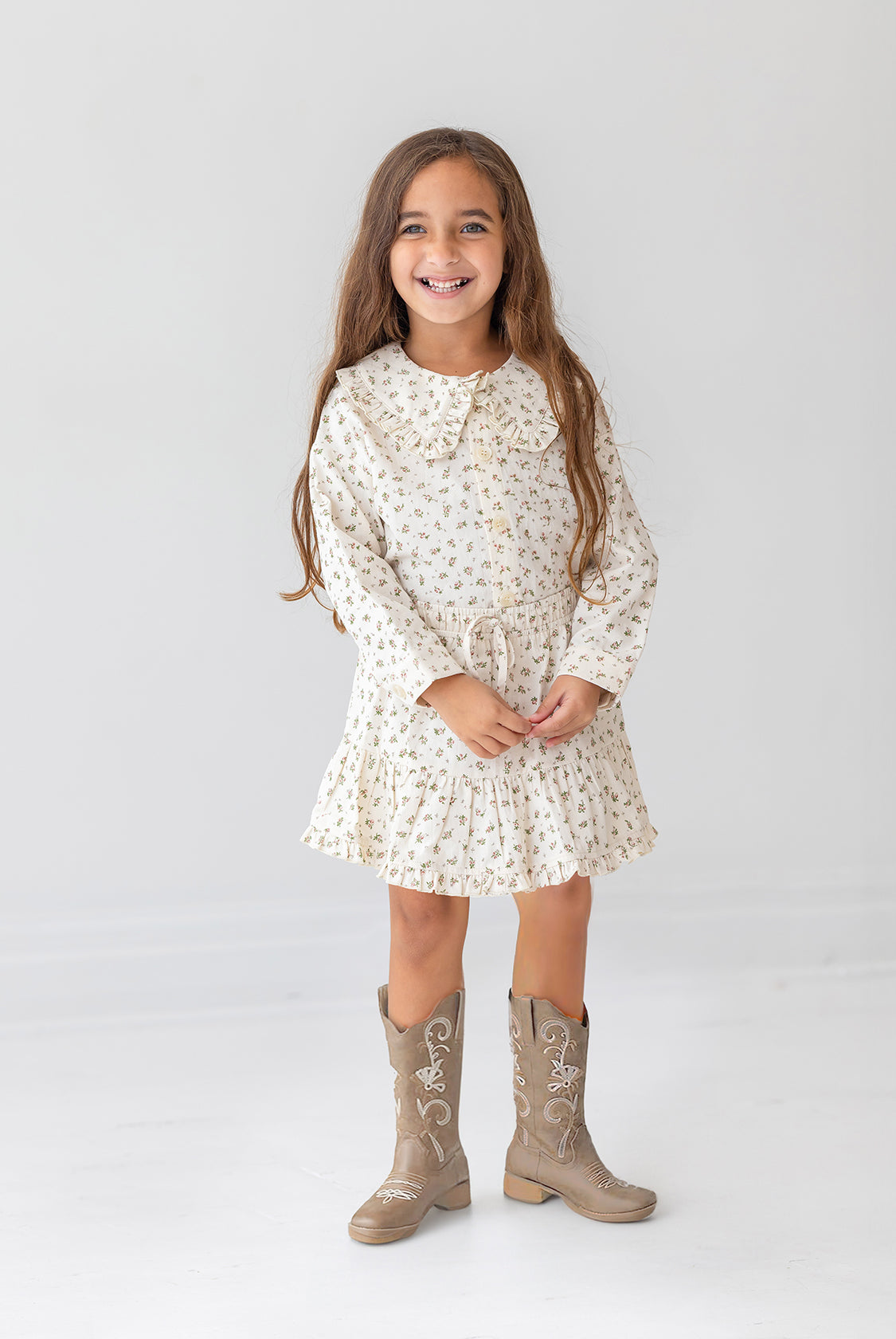 A young girl with long brown hair smiles against a white background, wearing the Mabel and Honey Juliette Two Piece Set and tan cowboy boots.