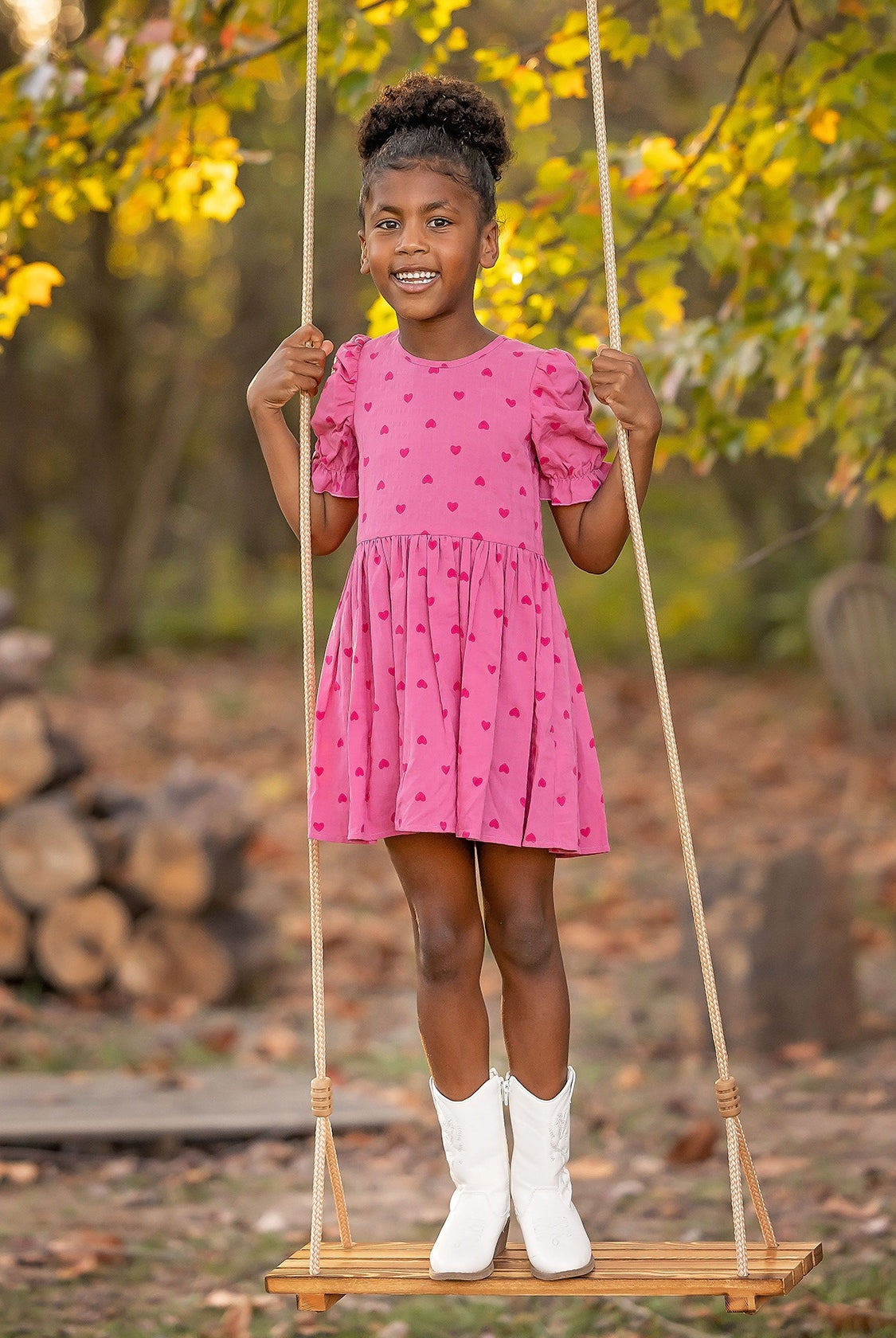 A young girl beams on a wooden swing outdoors, wearing the Mabel and Honey Perfect in Pink Hearts Short Sleeve Dress, paired with white boots amid autumn trees and stacked firewood.
