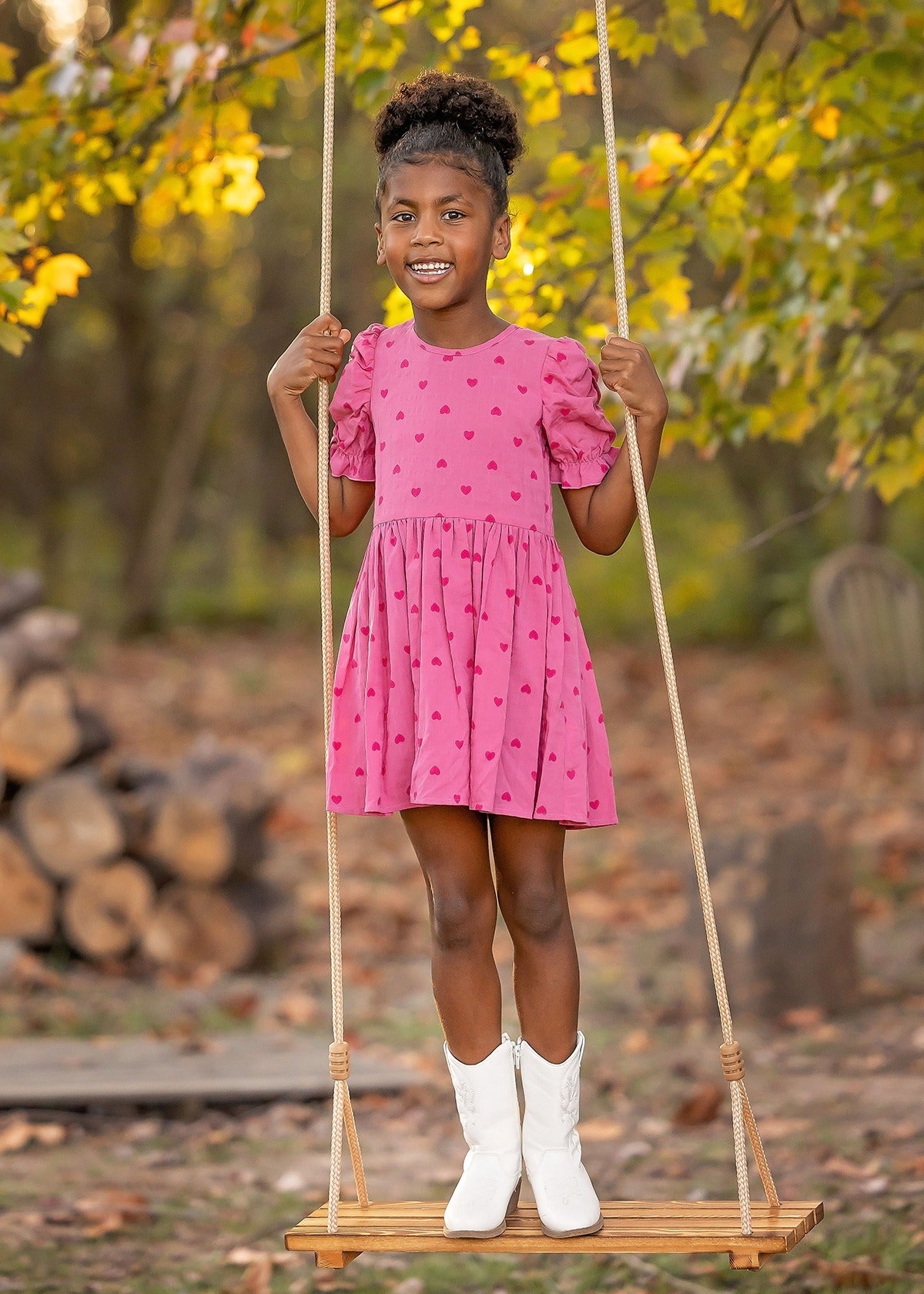A young girl beams on a wooden swing outdoors, wearing the Mabel and Honey Perfect in Pink Hearts Short Sleeve Dress, paired with white boots amid autumn trees and stacked firewood.