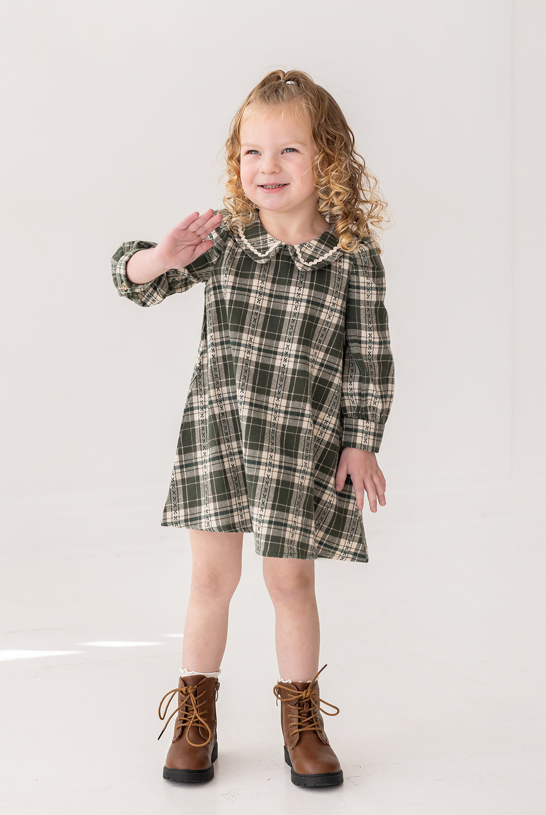 A young girl with curly blonde hair smiles and waves while modeling the Mabel and Honey Plaid & Pine Dress against a plain white background.