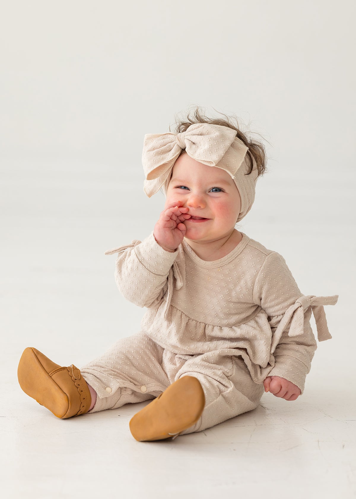 A smiling baby girl in a Mabel and Honey Cinnamon Sugar Romper with bow details and a matching headband sits on the floor, touching her mouth while wearing tan shoes against a plain white background.