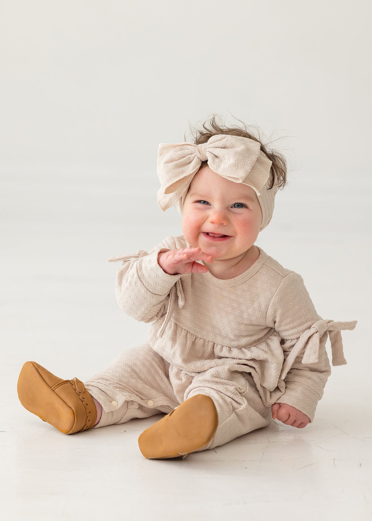 A smiling baby girl sits on a white floor against a plain background, wearing the Mabel and Honey Cinnamon Sugar Romper with bow-detailed sleeves, a matching large bow headband, and brown shoes.