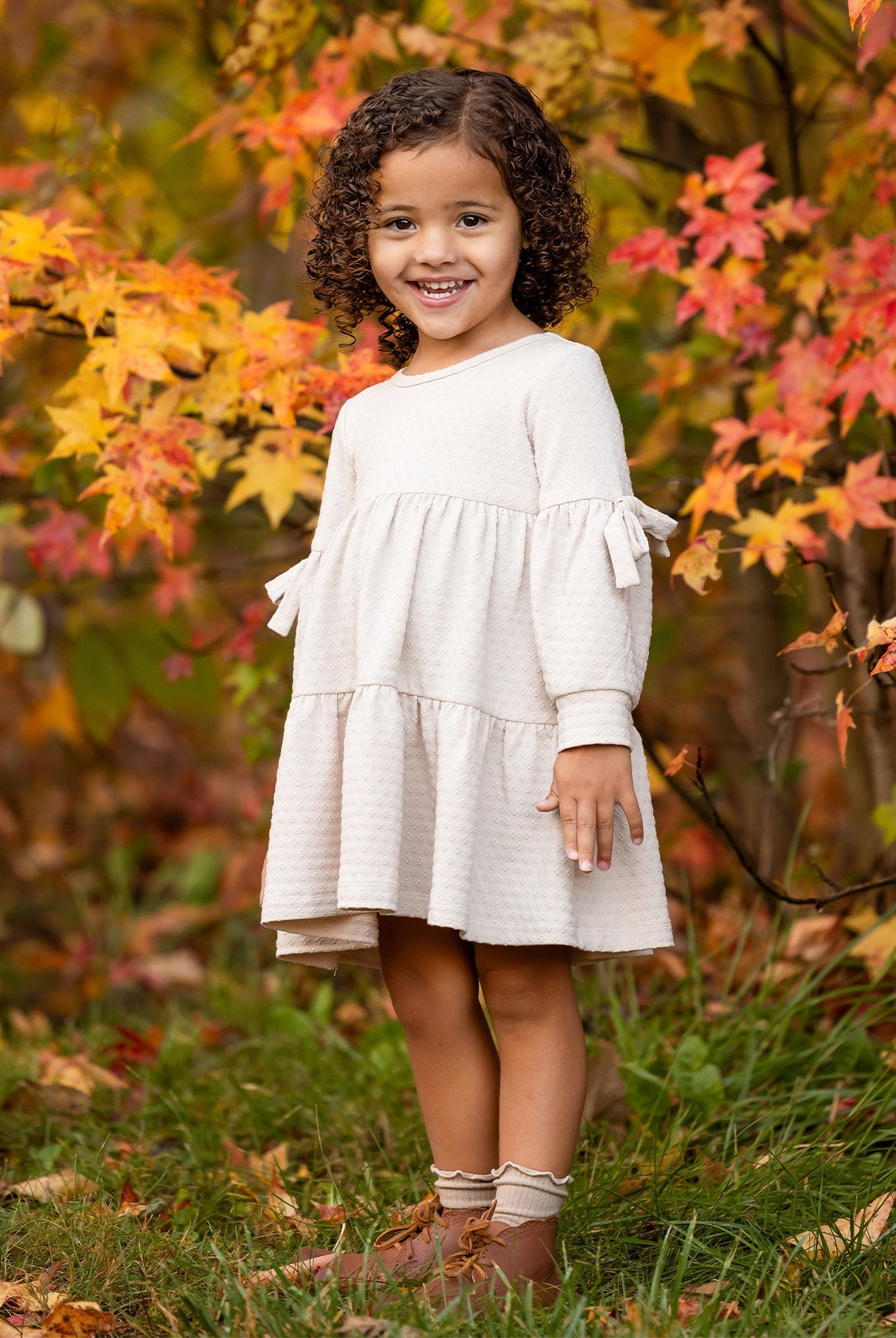 A young girl with curly hair smiles outdoors on green grass, wearing the Mabel and Honey Cinnamon Sugar Dress with a tiered silhouette and brown boots, standing in front of autumn trees with vibrant orange and yellow leaves.