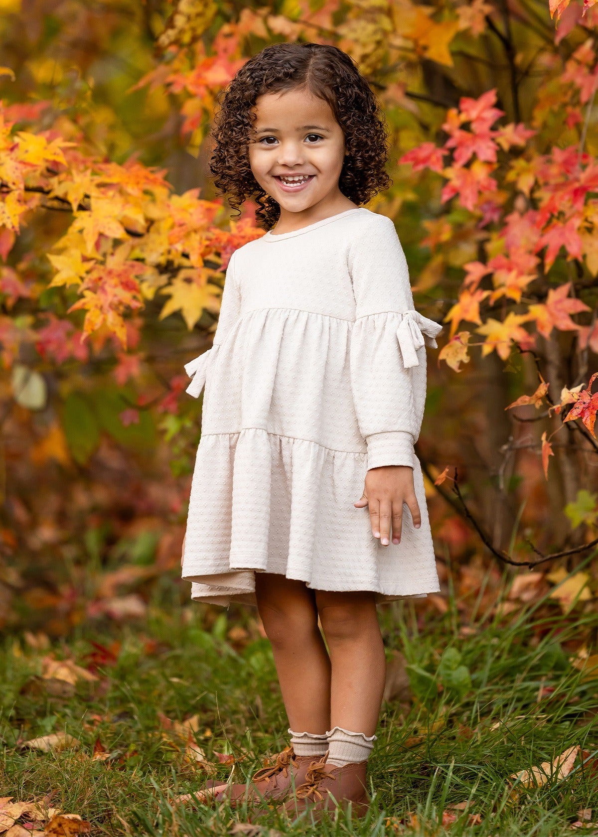 A young girl with curly hair smiles outdoors on green grass, wearing the Mabel and Honey Cinnamon Sugar Dress with a tiered silhouette and brown boots, standing in front of autumn trees with vibrant orange and yellow leaves.
