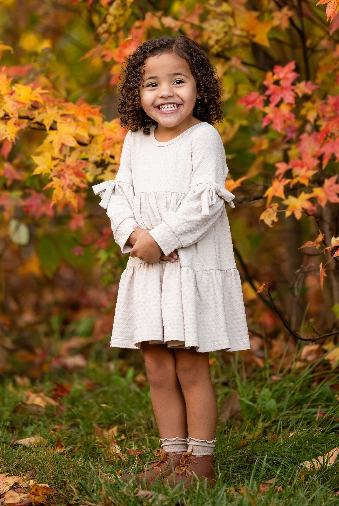 A young girl with curly hair smiles outdoors amid autumn leaves, wearing the Mabel and Honey Cinnamon Sugar Dress with sleeve bows and a tiered silhouette, paired with brown boots as she stands on green grass.