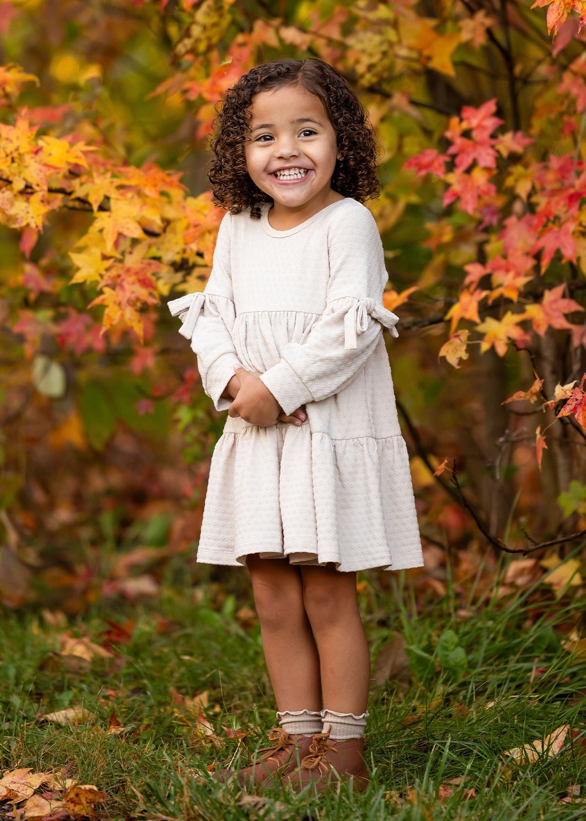 A young girl with curly hair smiles outdoors amid autumn leaves, wearing the Mabel and Honey Cinnamon Sugar Dress with sleeve bows and a tiered silhouette, paired with brown boots as she stands on green grass.