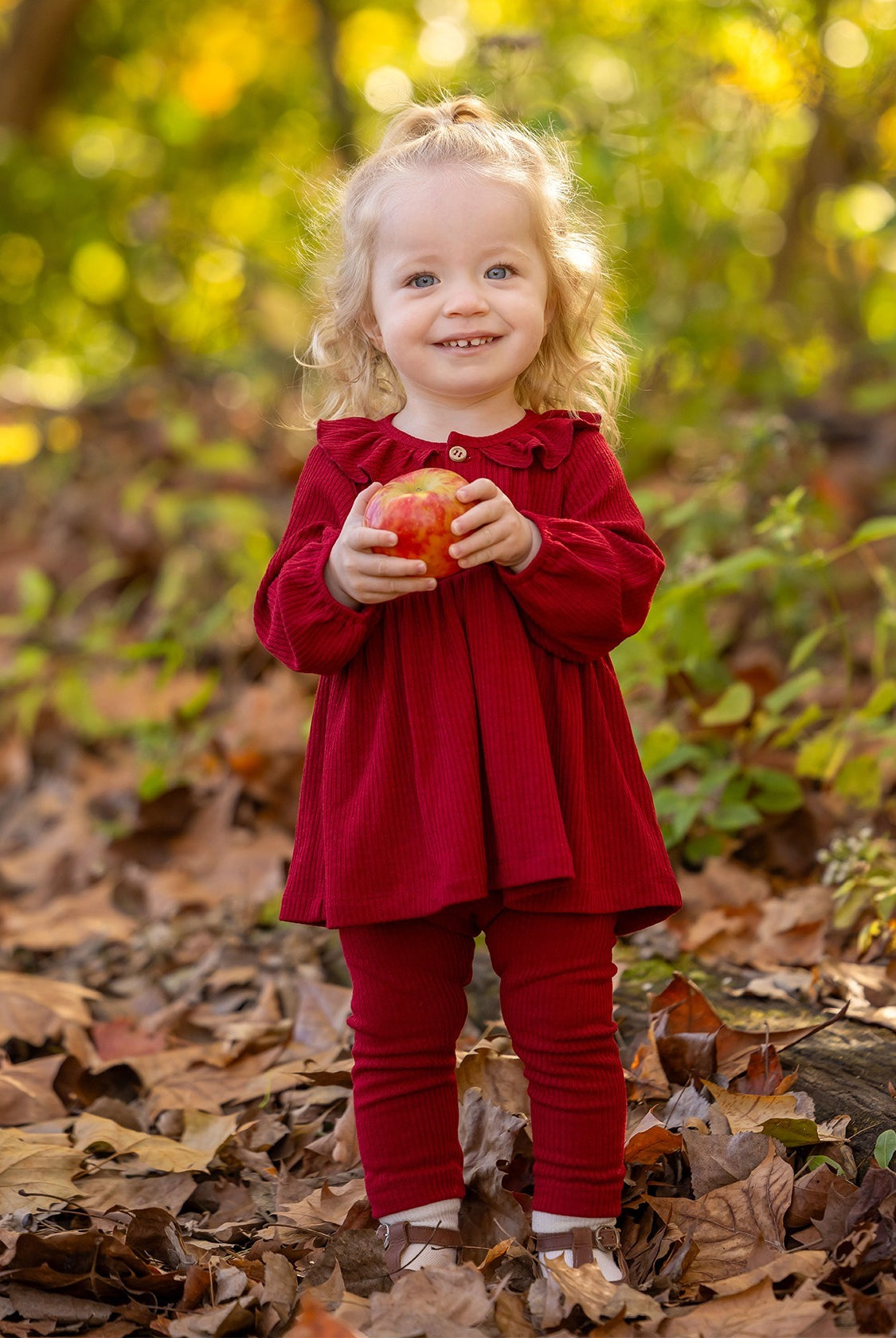 A smiling toddler with curly blonde hair stands outdoors on fallen leaves, wearing the Mabel and Honey Rosie Two Piece Set with ribbed leggings and a ruffled collar, holding a red apple against a blurred green and yellow foliage background.
