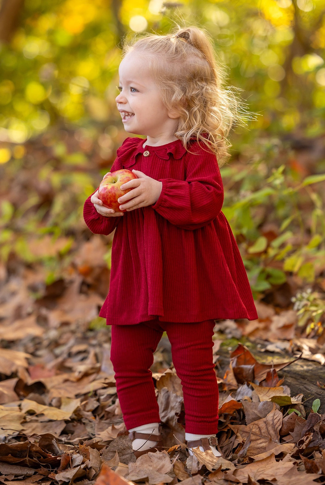 A smiling toddler in the Rosie Two Piece Set by Mabel and Honey stands among autumn leaves outdoors, holding a red apple with both hands. Green and yellow foliage is visible in the background.