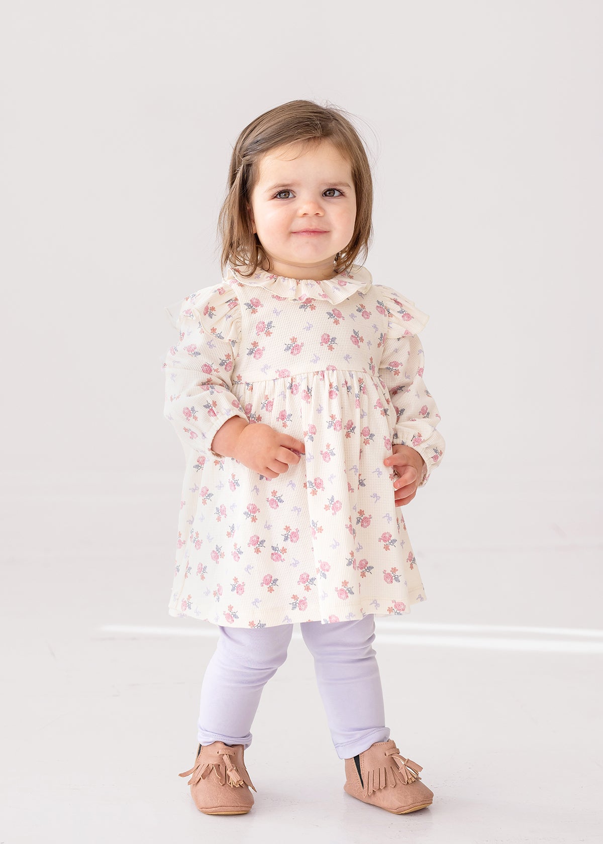 A young toddler stands indoors, smiling at the camera in the Mia Two Piece Set by Mabel and Honey—an adorable girls’ outfit featuring a cream ruffled blouse with pink floral patterns and light purple leggings.