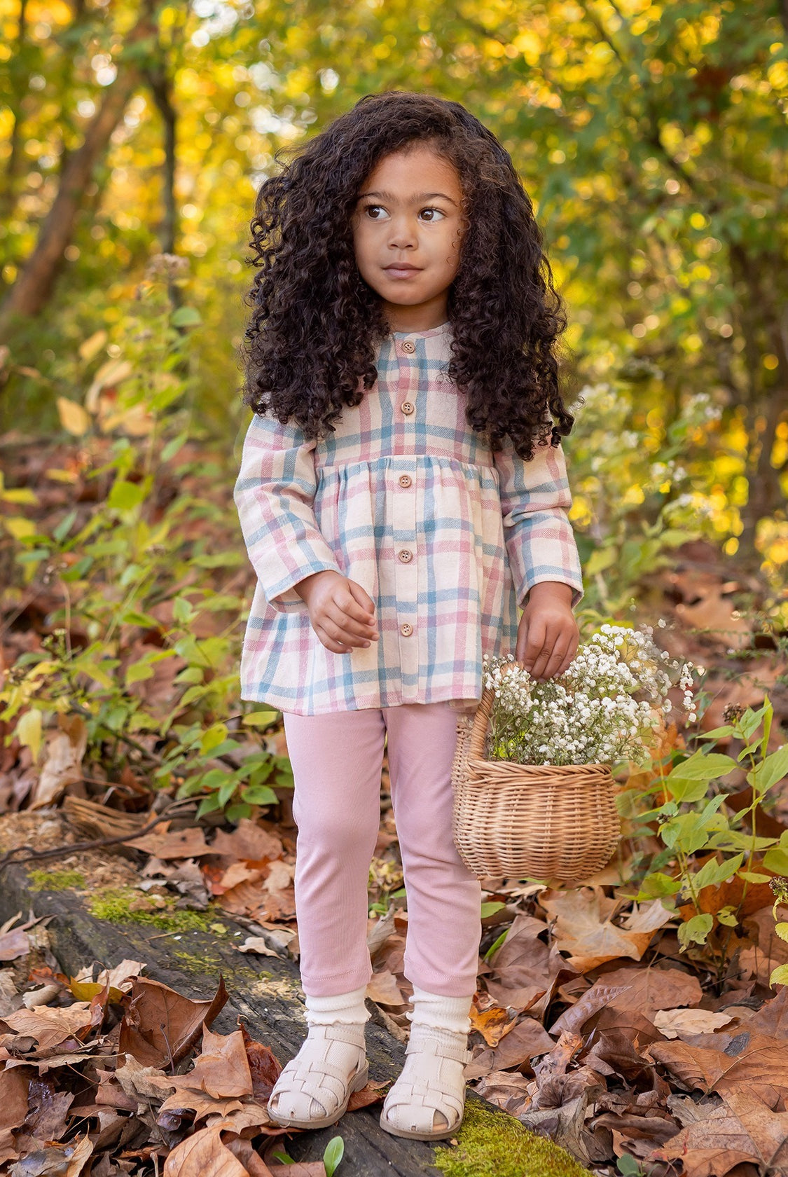 A young girl stands outdoors on fallen leaves, wearing the Phoebe Two Piece Set by Mabel and Honey. She holds a wicker basket of white flowers, with greenery and trees in the background.