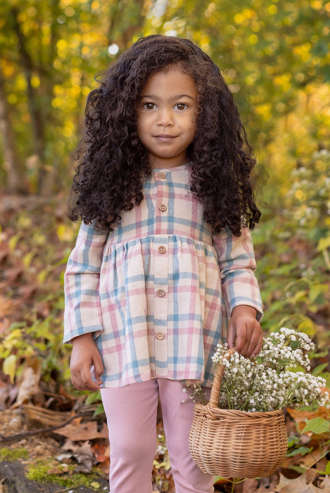 A young girl with long curly hair stands in a forest, wearing the Phoebe Two Piece Set by Mabel and Honey—a plaid top and pink leggings. She holds a basket of wildflowers and looks at the camera, surrounded by autumn leaves and greenery.