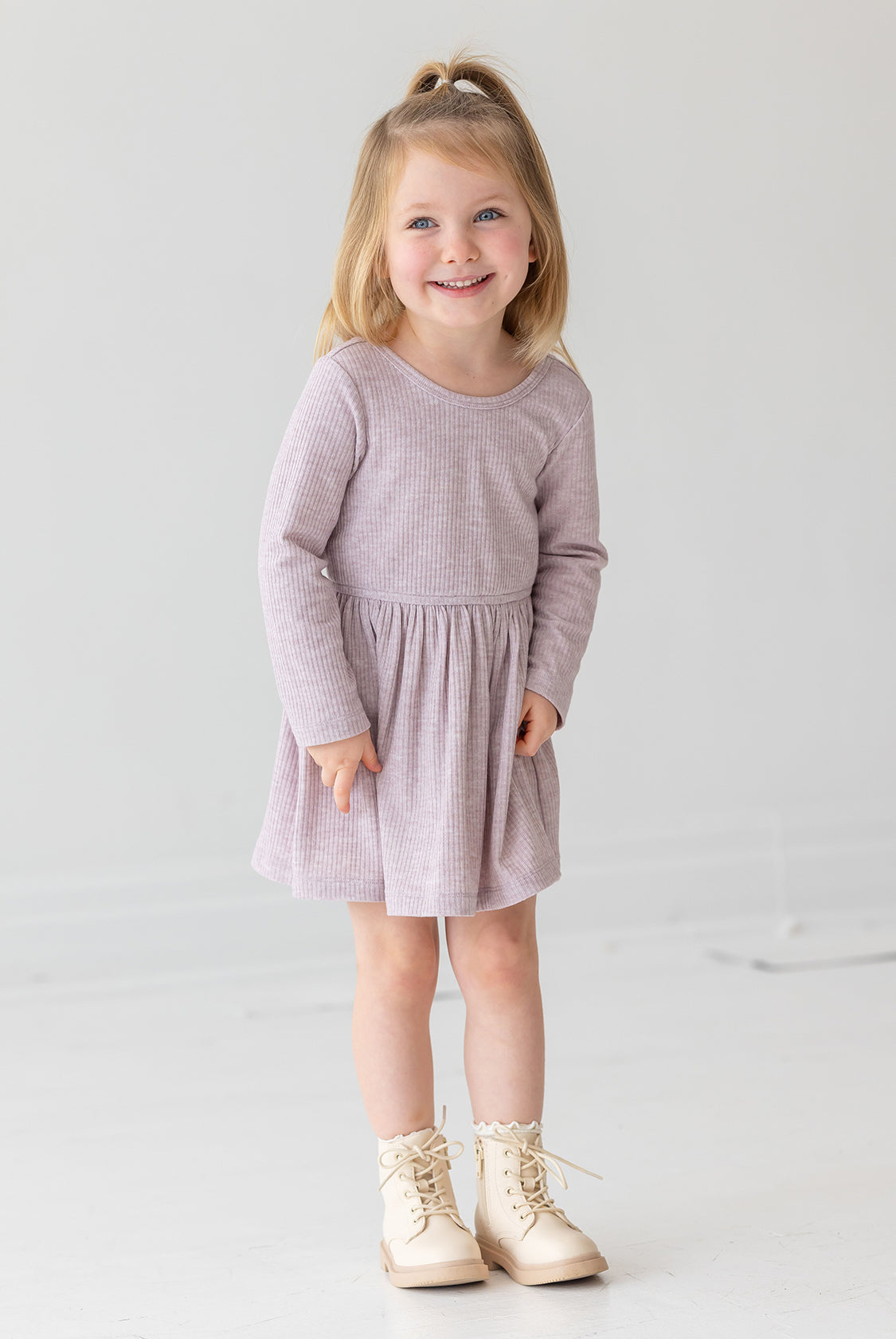 A young girl with blonde hair in a half ponytail smiles while standing against a plain white background. She wears the Heather Dress by Mabel and Honey, a light purple long-sleeve dress with a gathered skirt, paired with cream lace-up boots.