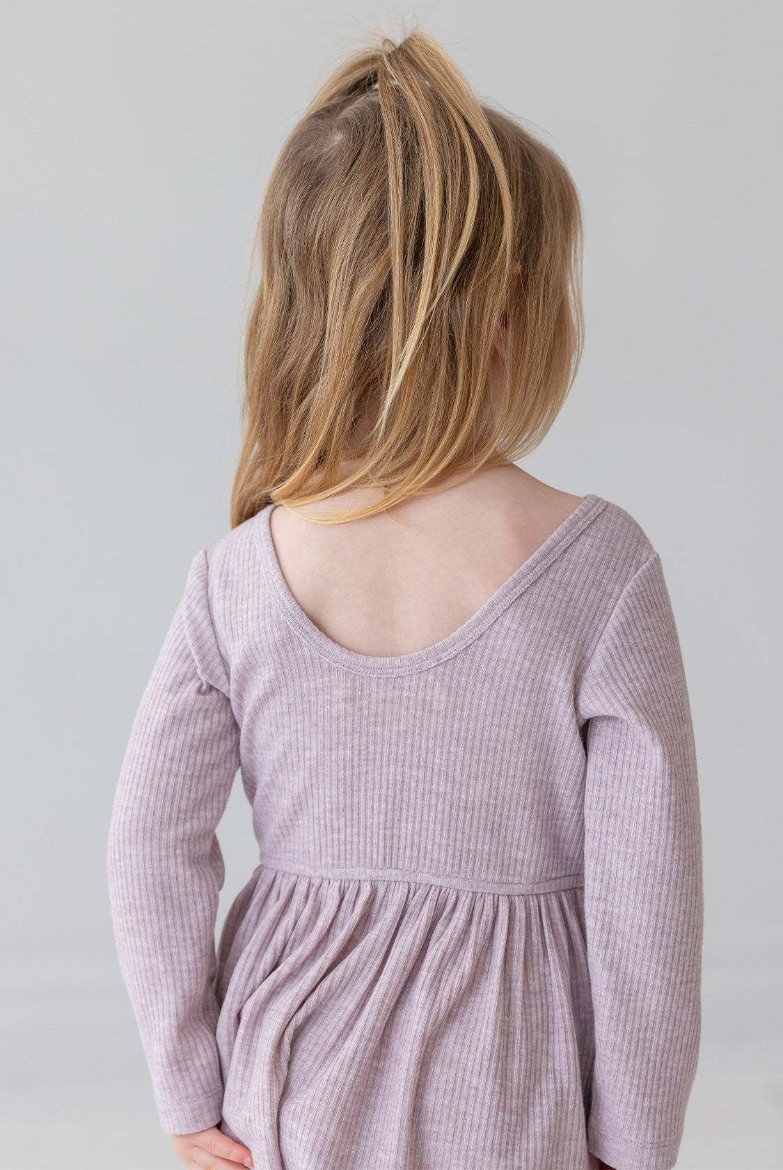 A young child with light brown hair in a half ponytail wears the Mabel and Honey Heather Dress, a long sleeve ribbed pink dress with a gathered skirt, standing and facing away from the camera against a plain light background.