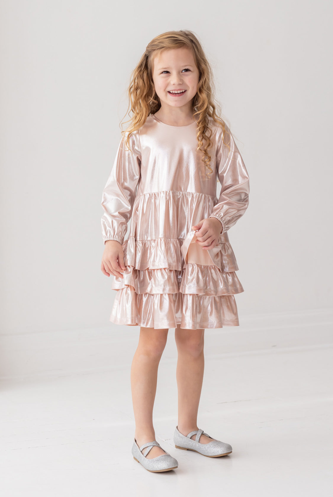 A smiling young girl with long, curly blonde hair wears the Isobella and Chloe Golden Glow Long Sleeve Dress with light pink ruffles and silver shoes, standing against a white background for a perfect special occasion look.
