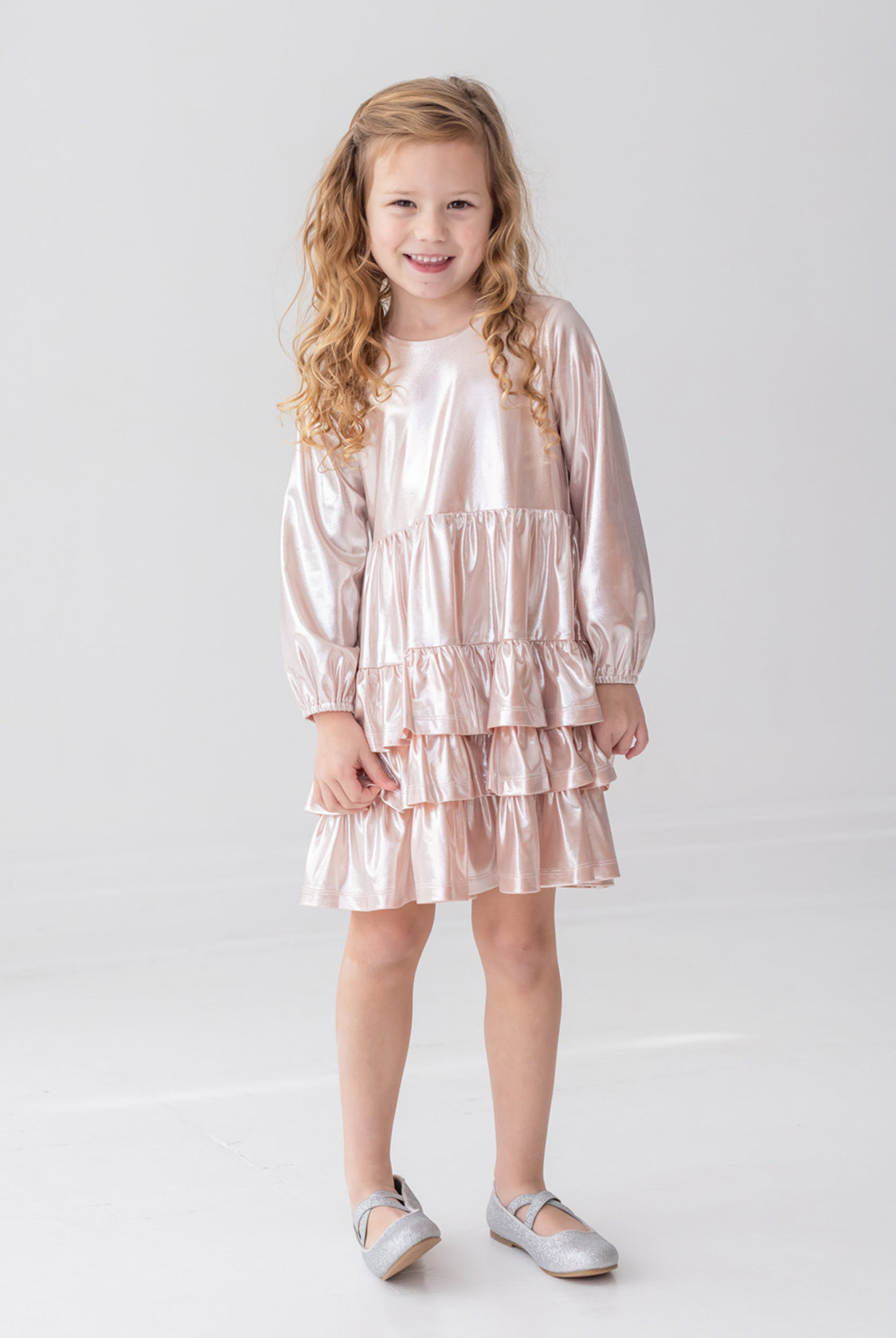 A young girl with long, curly blonde hair wears the Isobella and Chloe Golden Glow Long Sleeve Dress, standing and smiling in a bright white room—an ideal choice for any special occasion.