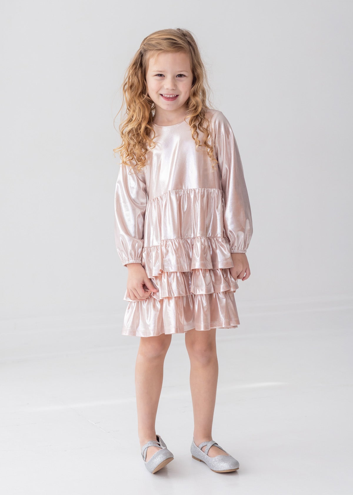 A young girl with long, curly blonde hair wears the Isobella and Chloe Golden Glow Long Sleeve Dress, standing and smiling in a bright white room—an ideal choice for any special occasion.