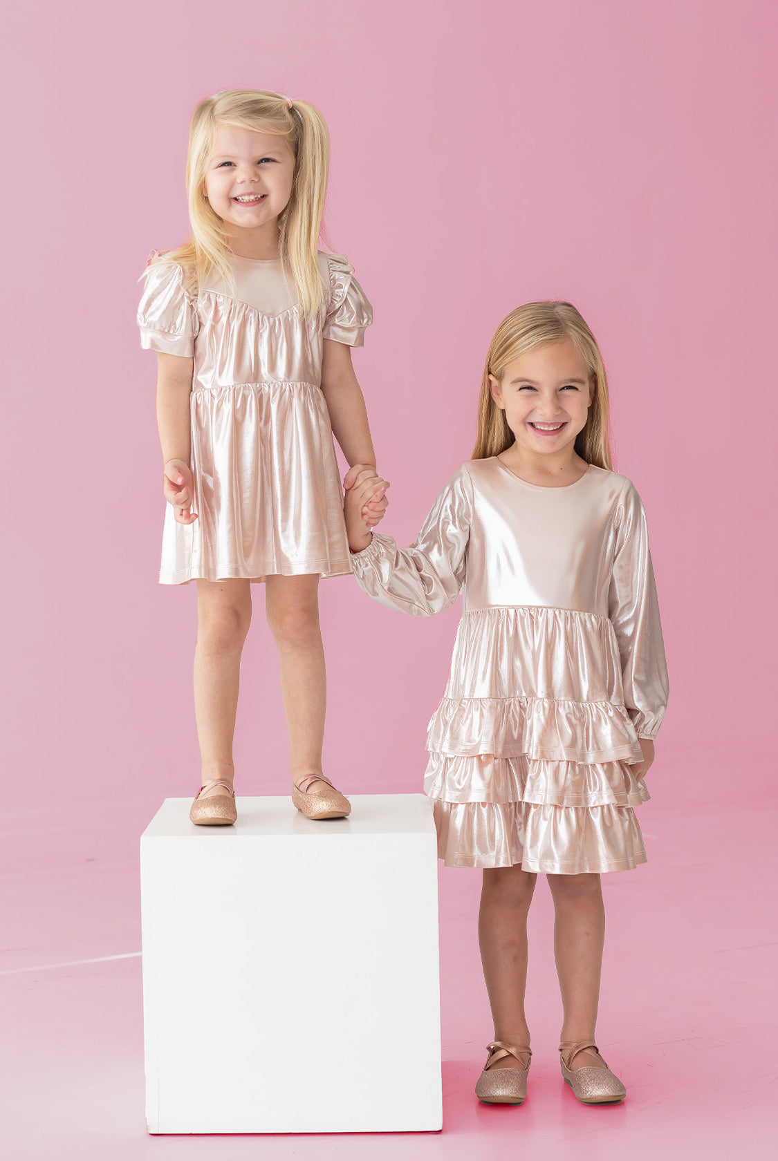 Two young girls in Isobella and Chloe's Golden Glow Long Sleeve Dresses smile against a pink background. One stands on a white cube, the other beside her. Holding hands, they shine in the perfect special occasion look.