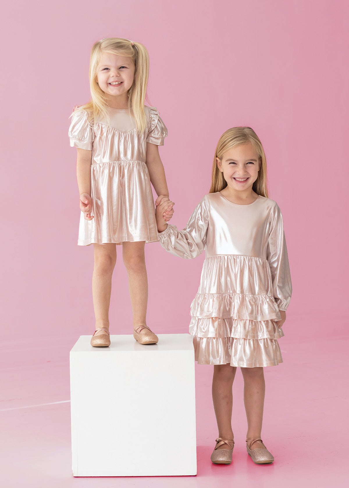 Two young girls in Isobella and Chloe's Golden Glow Long Sleeve Dresses smile against a pink background. One stands on a white cube, the other beside her. Holding hands, they shine in the perfect special occasion look.