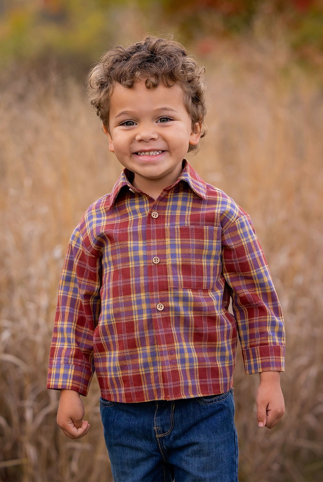 A young boy with curly hair smiles outdoors in tall grass, wearing the Beckett and Bear Noah Shirt—a red and yellow plaid cotton-polyester blend—paired with blue jeans. The blurred background features warm autumn colors.