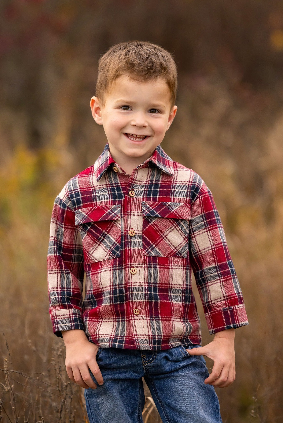 A young boy with short brown hair smiles outdoors, wearing the Beckett and Bear Henry Jacket. The background is softly blurred with brown and green tones, highlighting a natural setting.