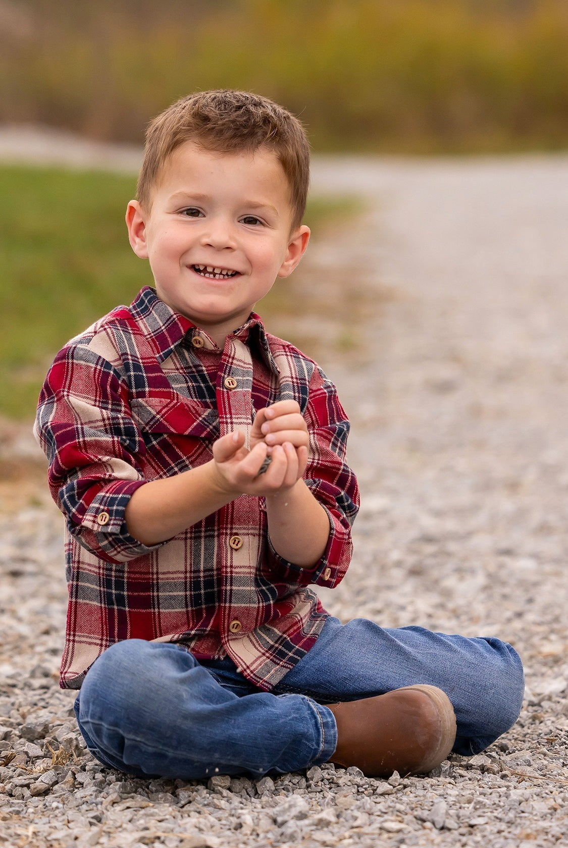 A young boy wearing the Beckett and Bear Henry Jacket sits cross-legged on a gravel path, smiling at the camera with small rocks in his hands, grass and blurred trees in the background.