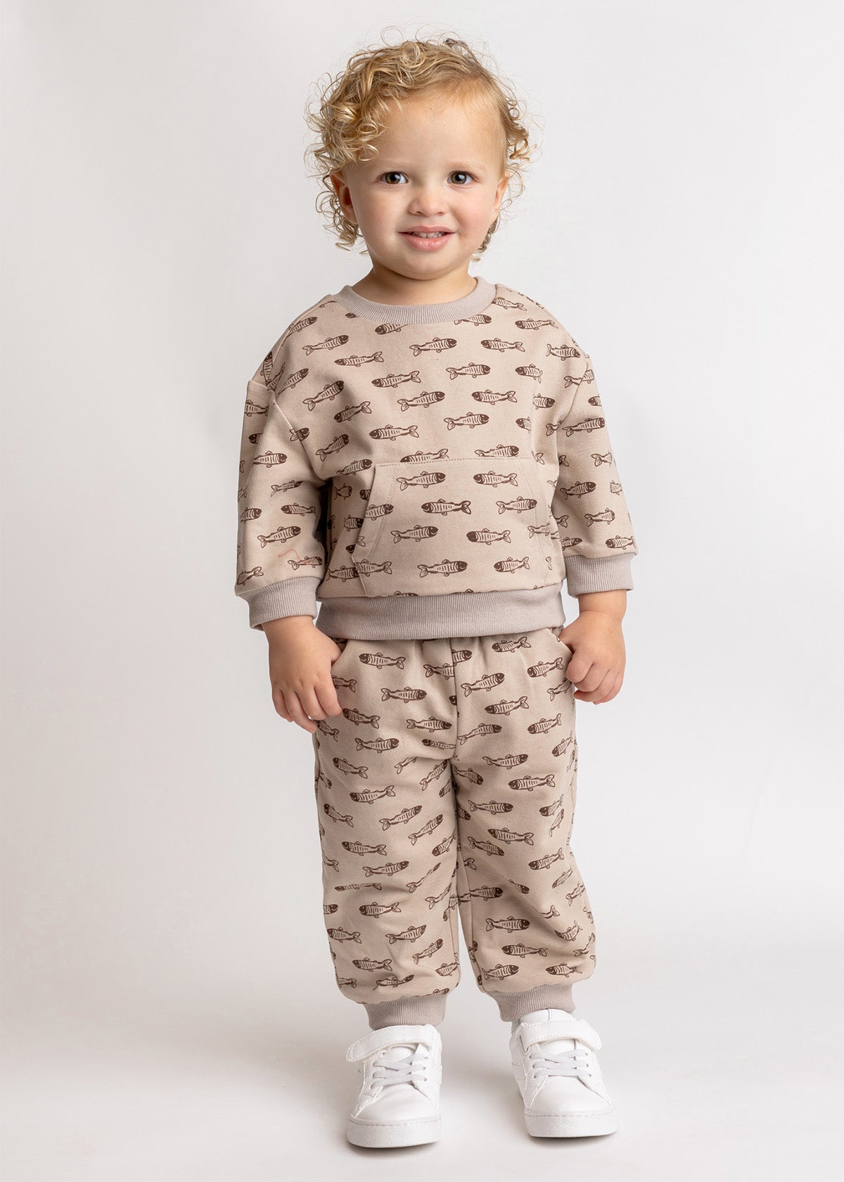 A young child with curly blond hair smiles in the Isaiah Two Piece Set by Beckett and Bear, featuring a beige kids sweatshirt and pants with brown cheetah print, paired with white socks and sneakers against a plain, light background.