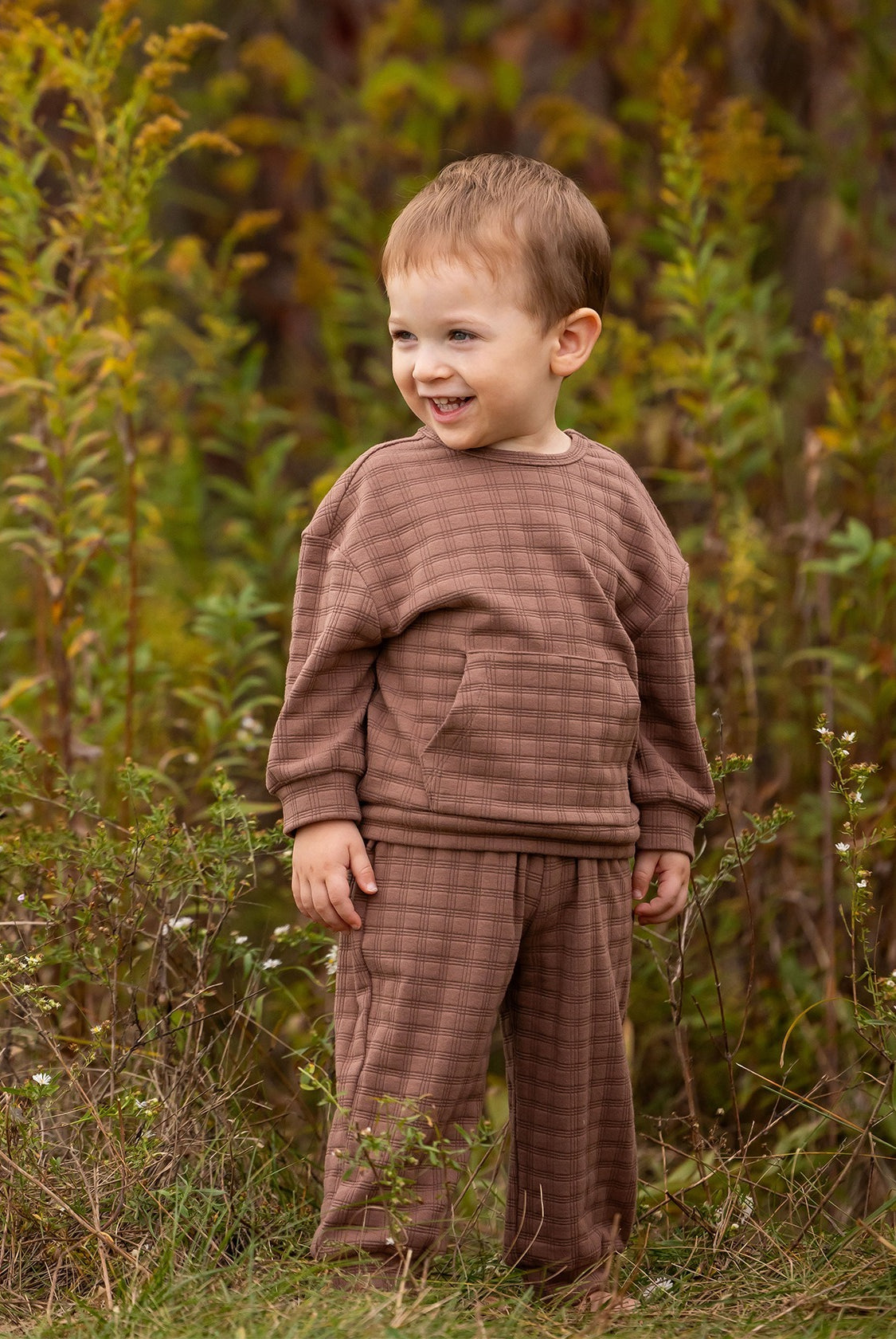A young child with short light brown hair wears the Theo Two Piece Set from Beckett and Bear, smiling while standing in a grassy outdoor area surrounded by tall green plants.