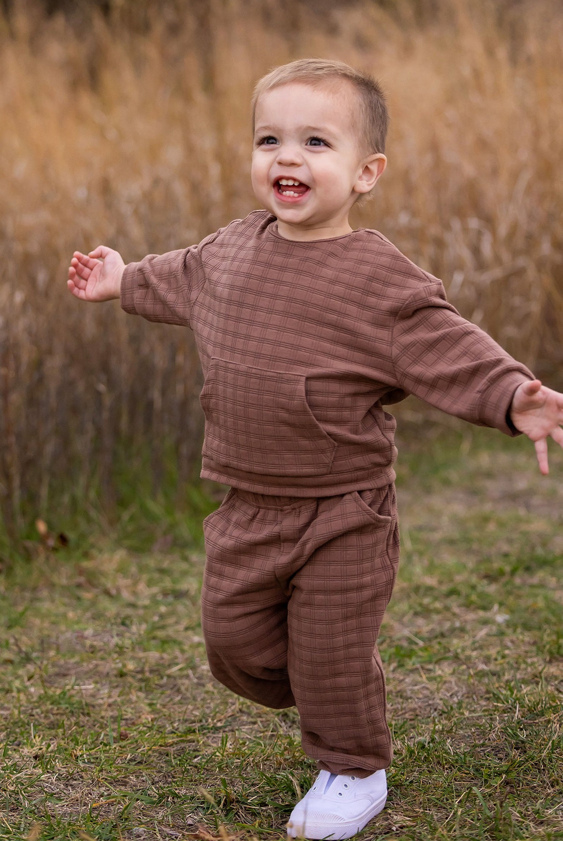 A young child wearing the Beckett and Bear Theo Two Piece Set and white sneakers walks with arms outstretched, smiling outdoors on grass with tall, dry grass in the background.