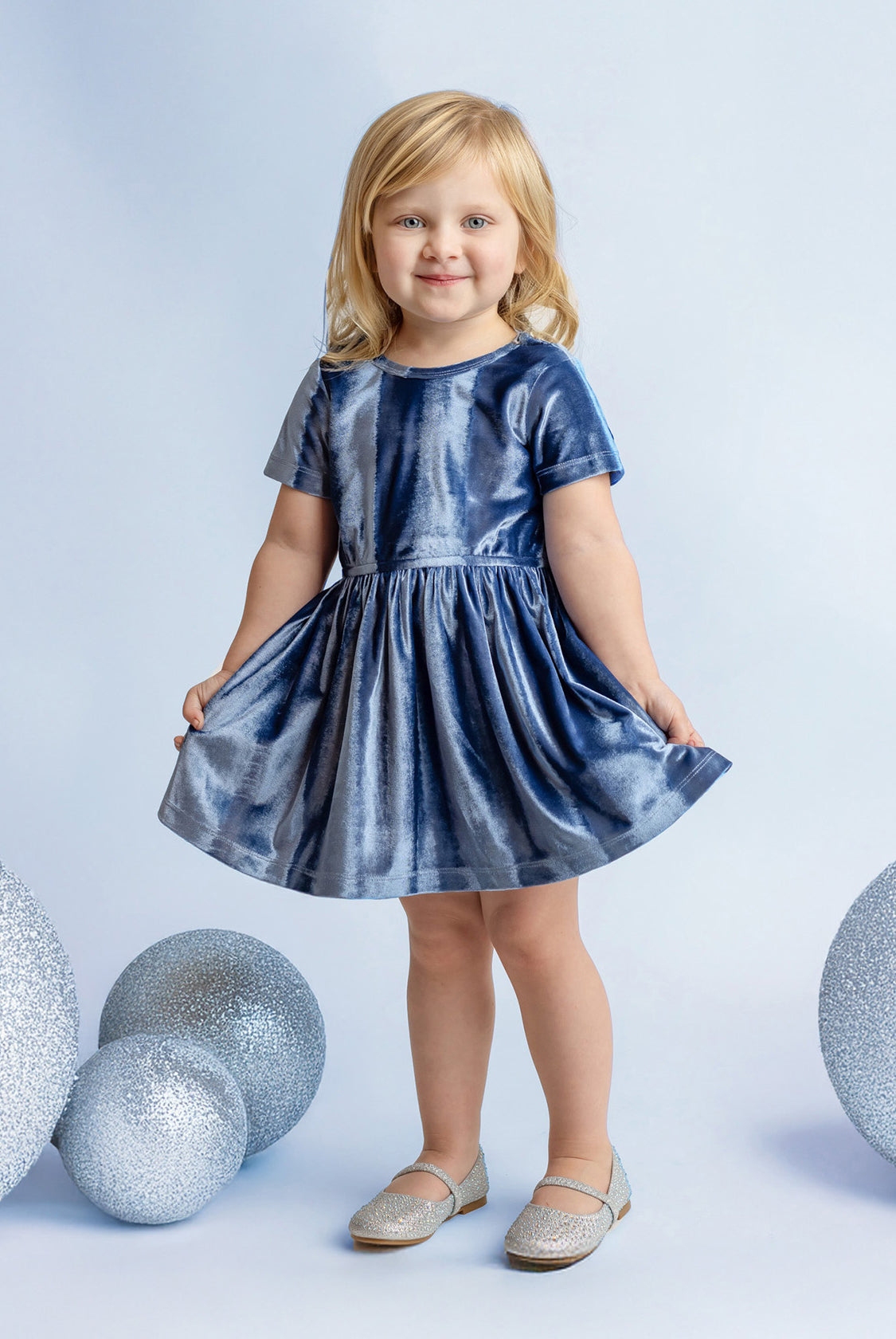 A young blonde girl smiles in the Isobella and Chloe Starry Blue Velvet Dress, holding the sides of her special occasion outfit, surrounded by large glittery silver spheres on a light blue background.