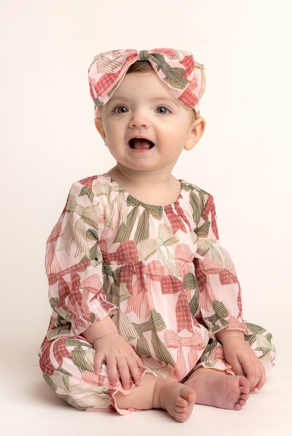 A baby smiles in Mabel and Honey’s Bow-tiful Romper—an adorable long-sleeve romper with a pink and green bow print, paired with a matching headband—set against a light, plain background.