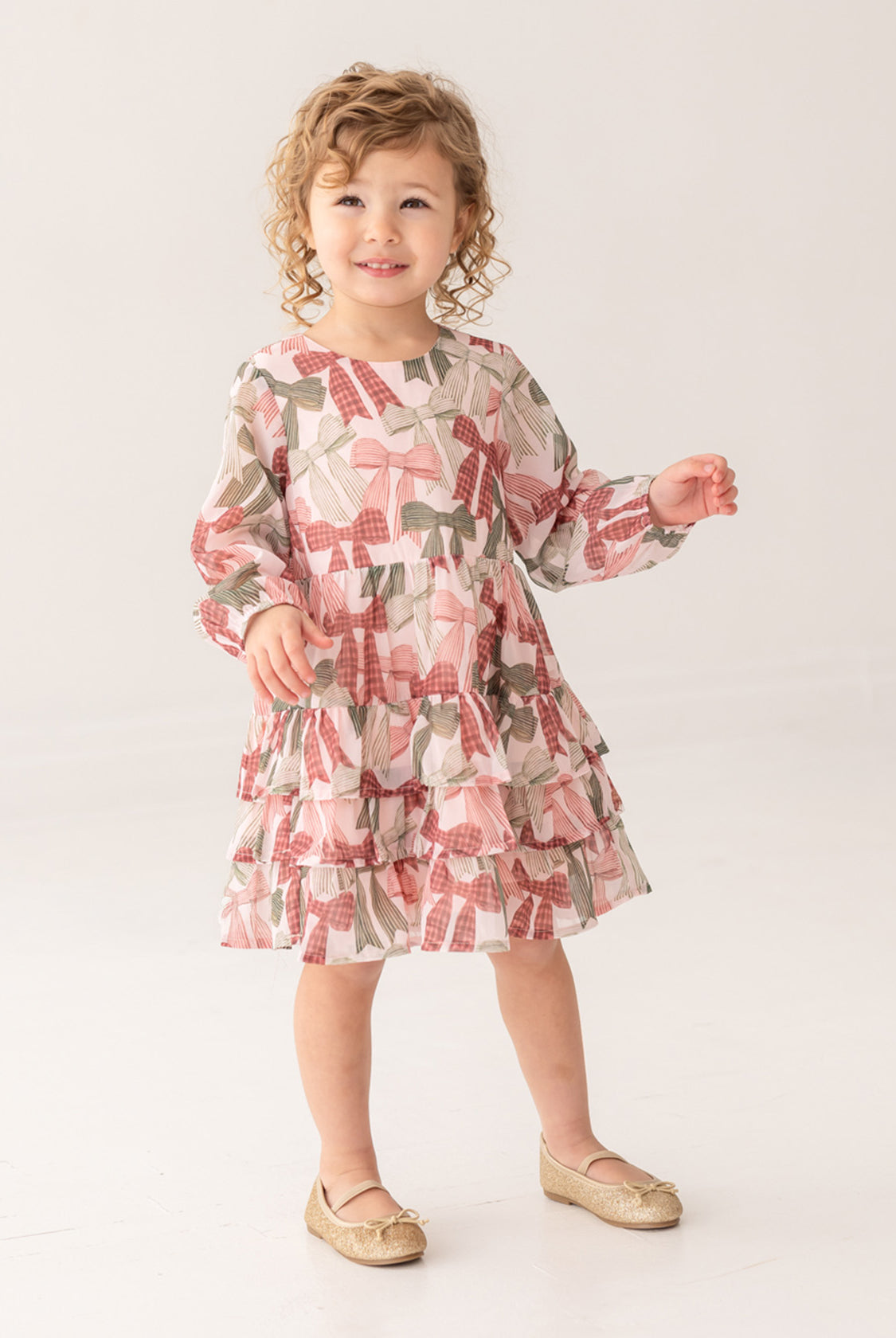 A young child with curly hair wears the Mabel and Honey Bow-tiful Dress, featuring a pink and green geometric pattern, paired with gold shoes, standing and smiling against a plain light background.